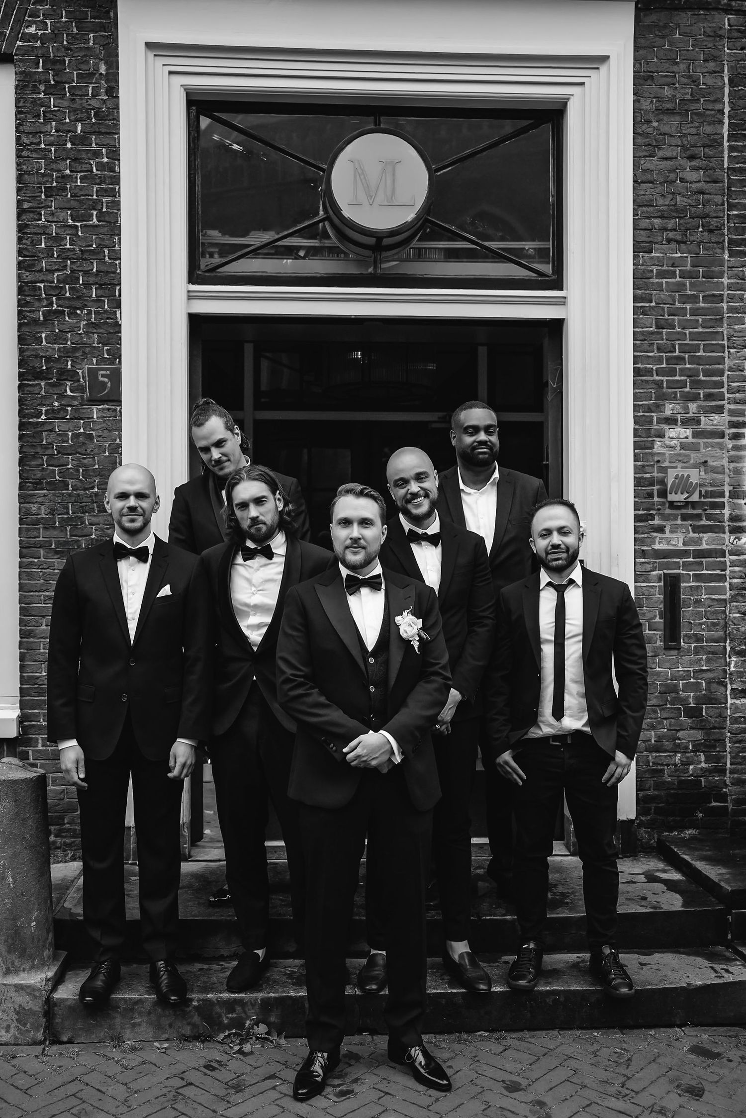 Group of groomsmen in formal attire posed on brick building steps.