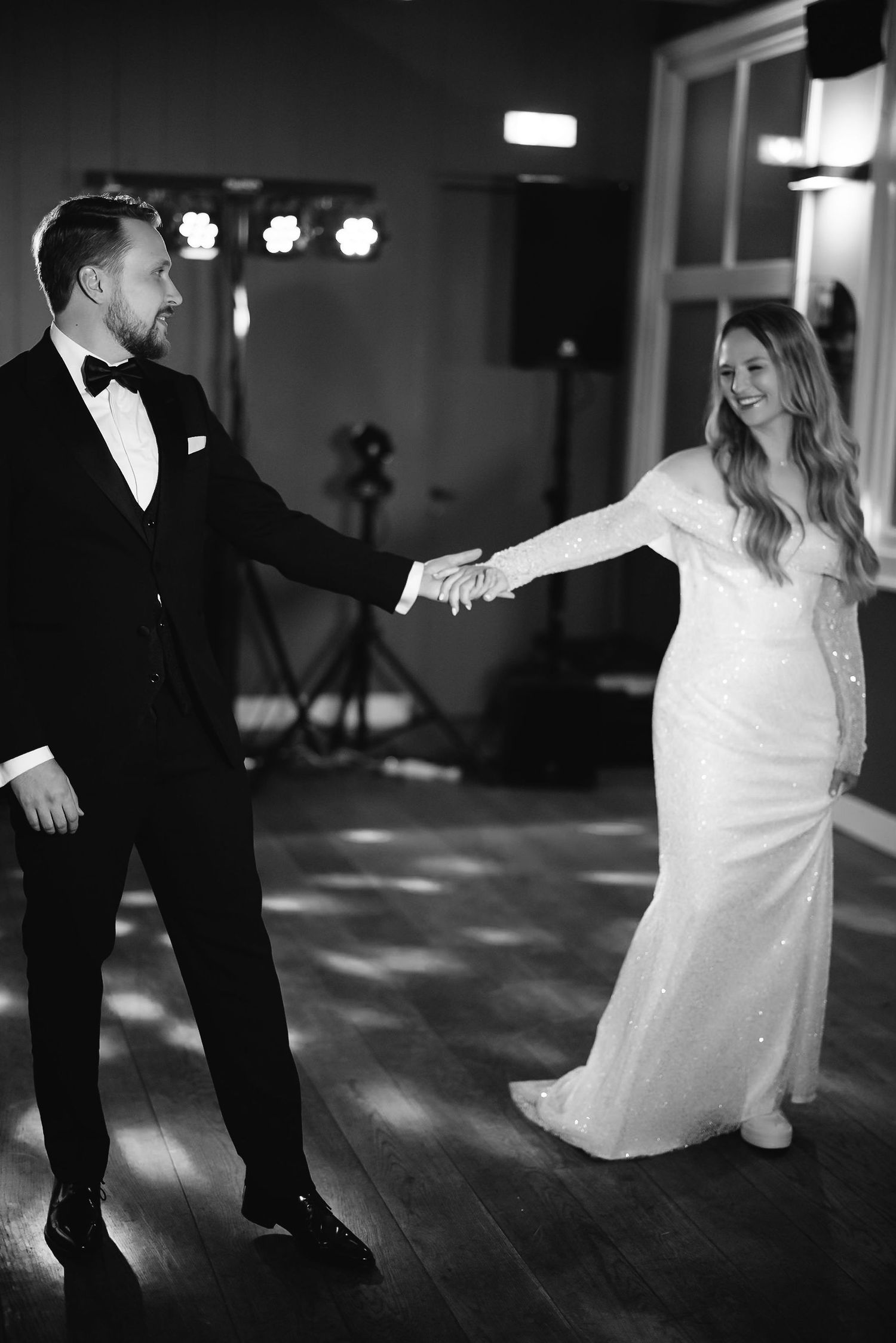 A couple shares their first dance in formal wedding attire on a wooden dance floor under mood lighting.