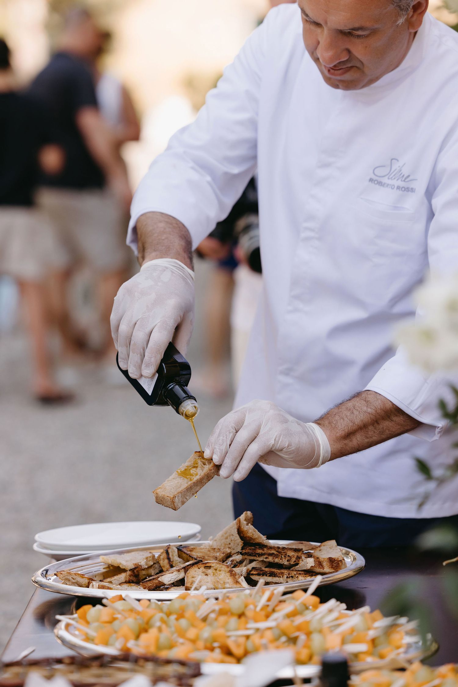 Chef in white uniform drizzling sauce over plated appetizers at catering event.