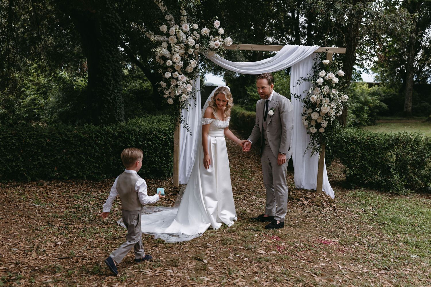 Intimate outdoor wedding ceremony under a white draped arch with floral decorations.