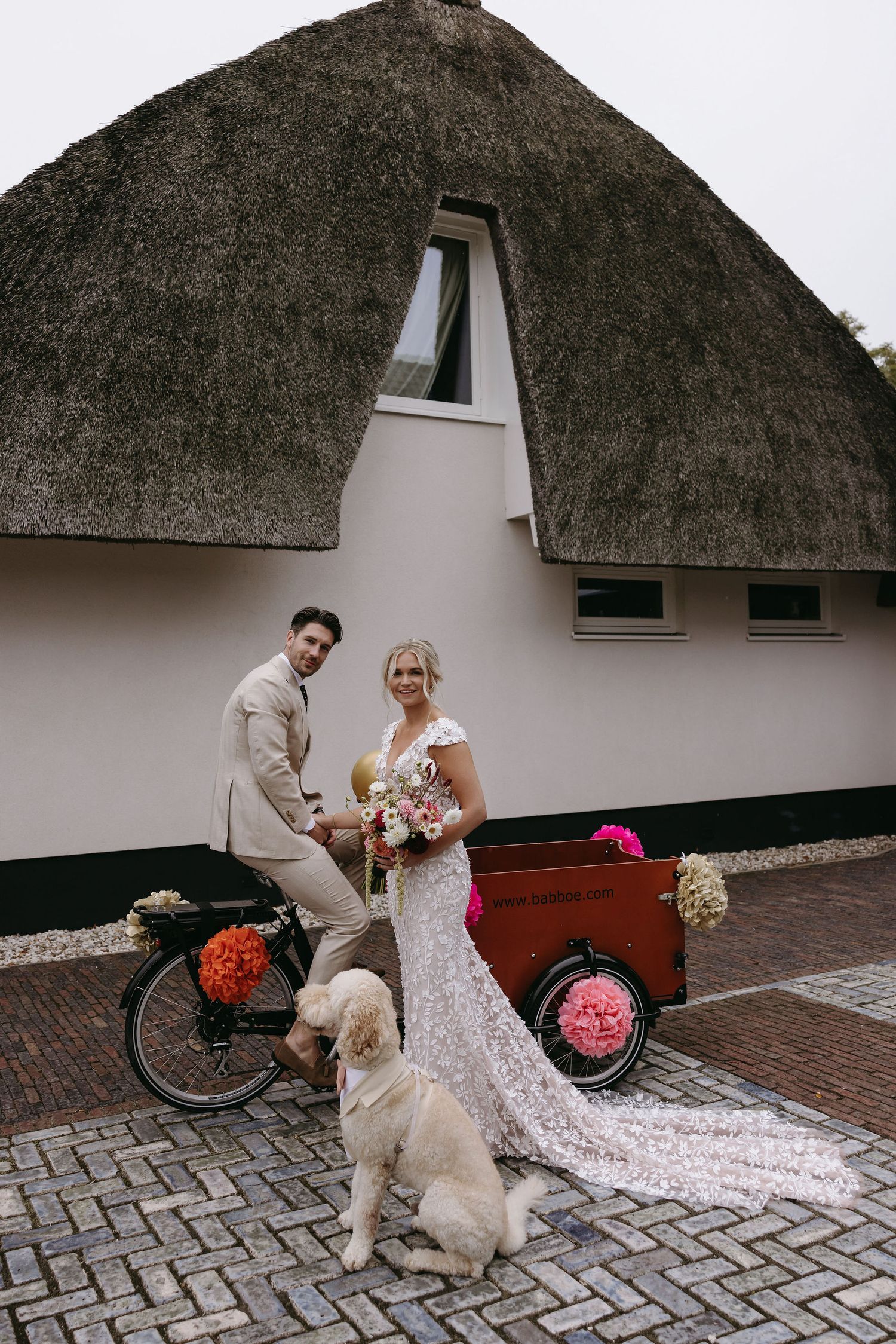 A couple poses with their white dog in front of a thatched-roof building with a vintage bicycle.