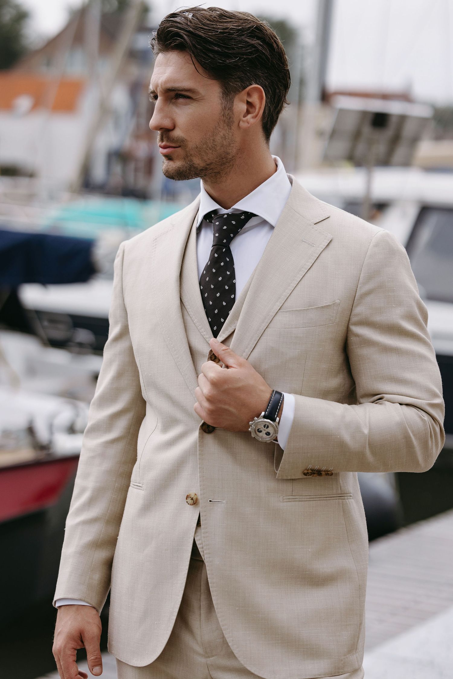 Elegant groom in beige suit and polka dot tie poses near marina.