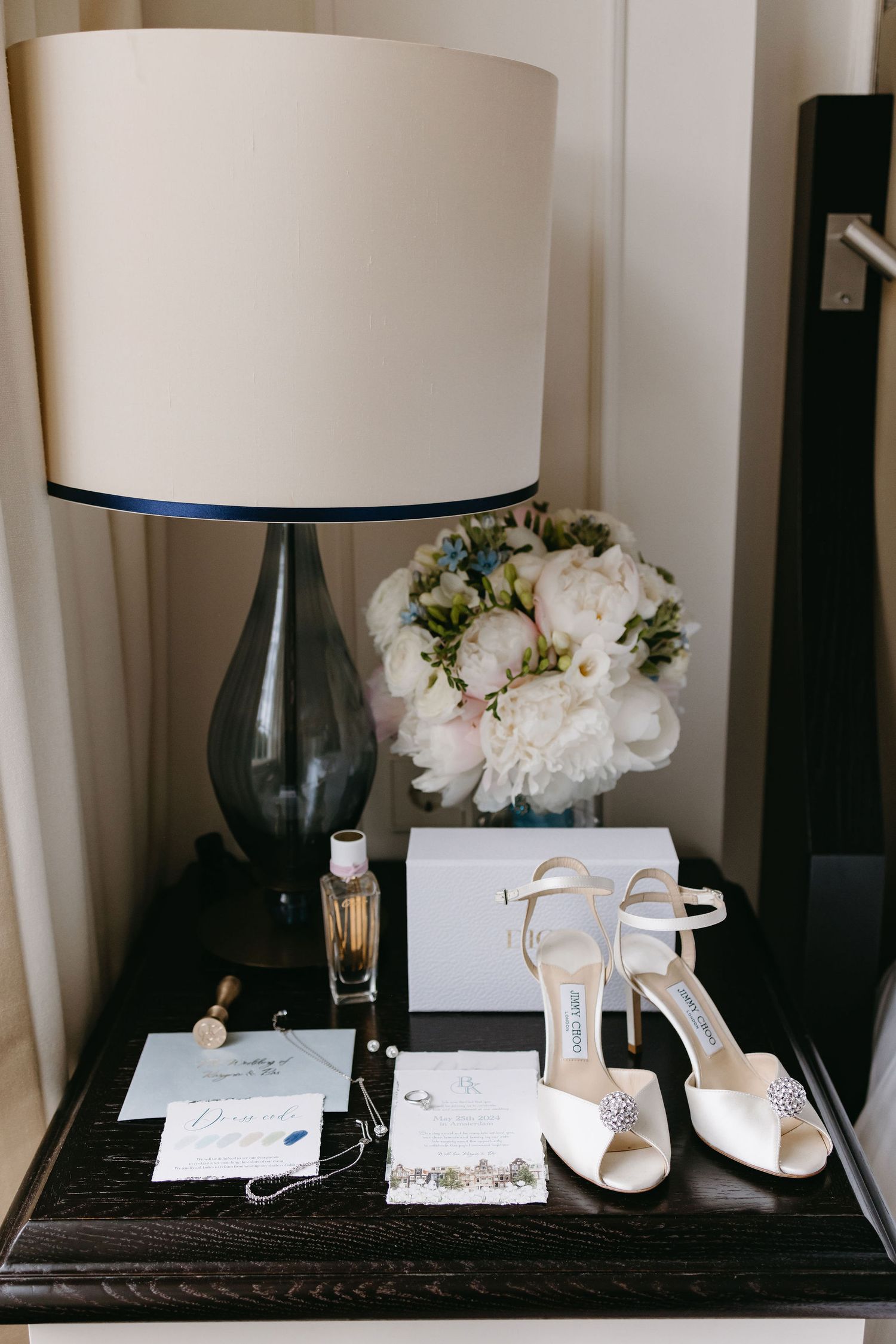 Wedding accessories including white shoes, jewelry and floral arrangements displayed on dark surface.