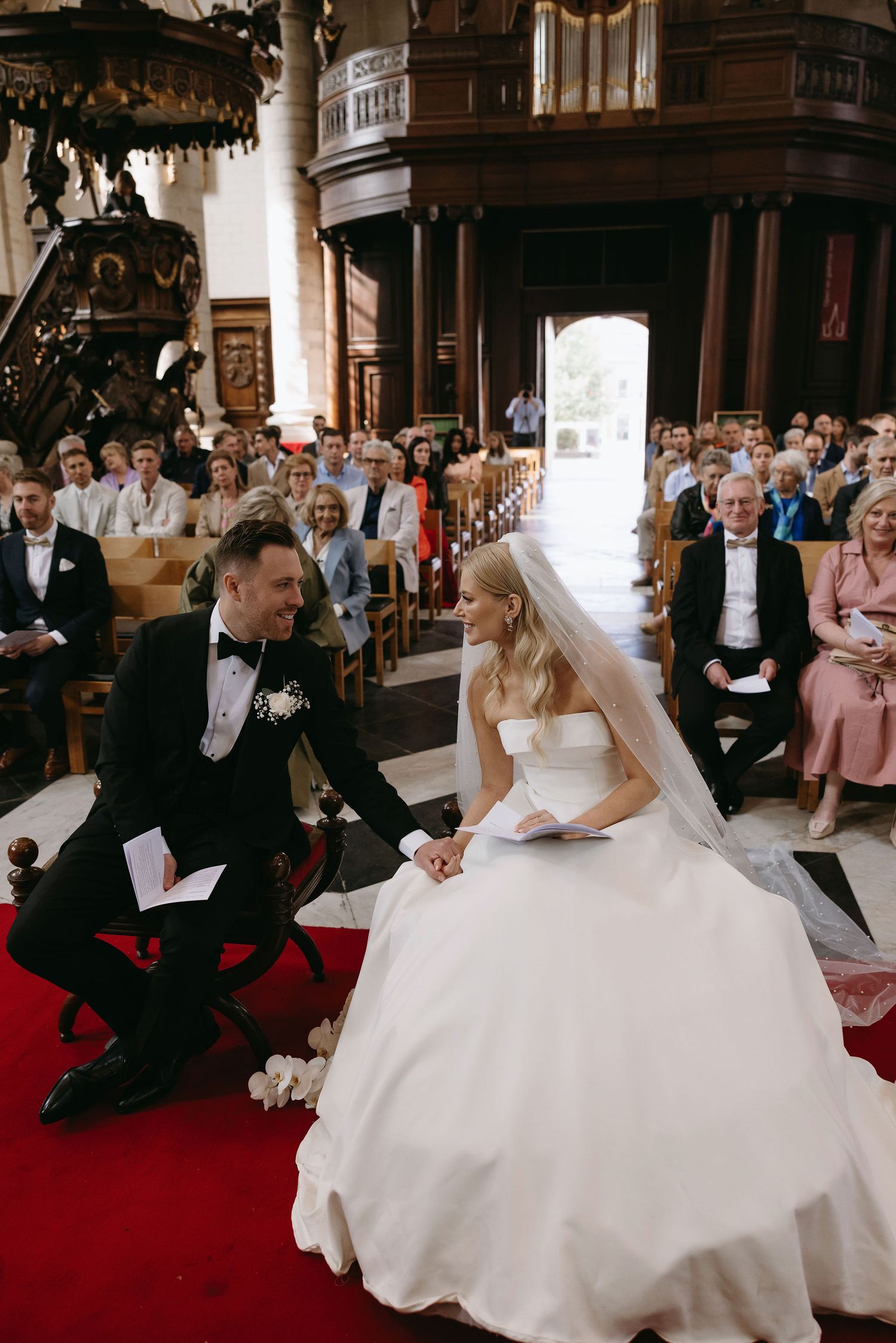 Wedding ceremony in an ornate church sanctuary with guests watching a couple seated on red carpet.