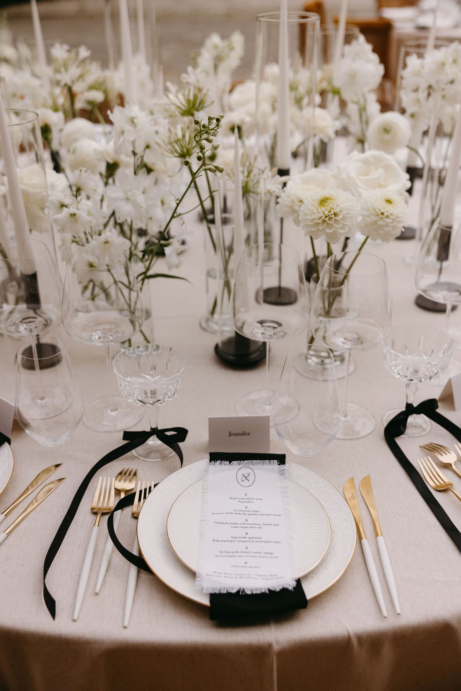 Elegant wedding table setting with white flowers in glass vases, gold cutlery and black ribbon accents.