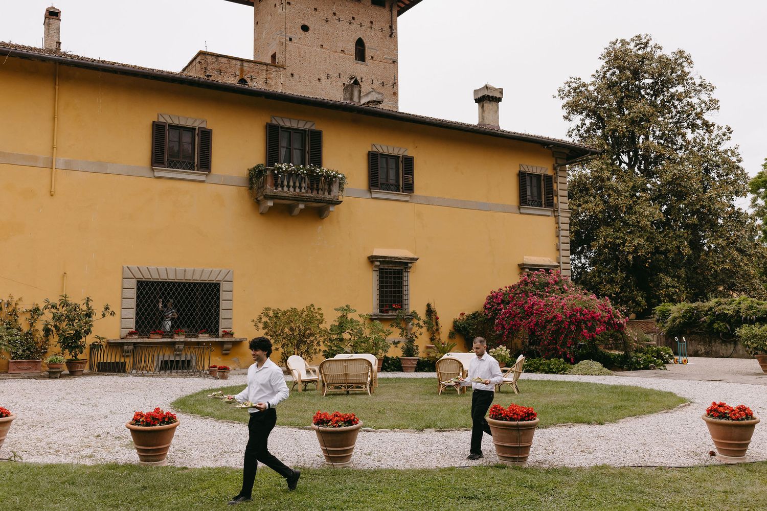 Staff member carrying items across courtyard of yellow Italian villa with terracotta planters.