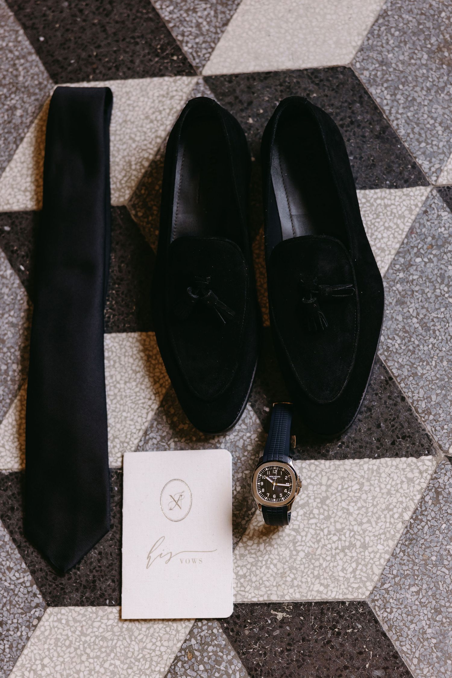 Black formal accessories including loafers, tie, watch and invitation on geometric patterned floor.