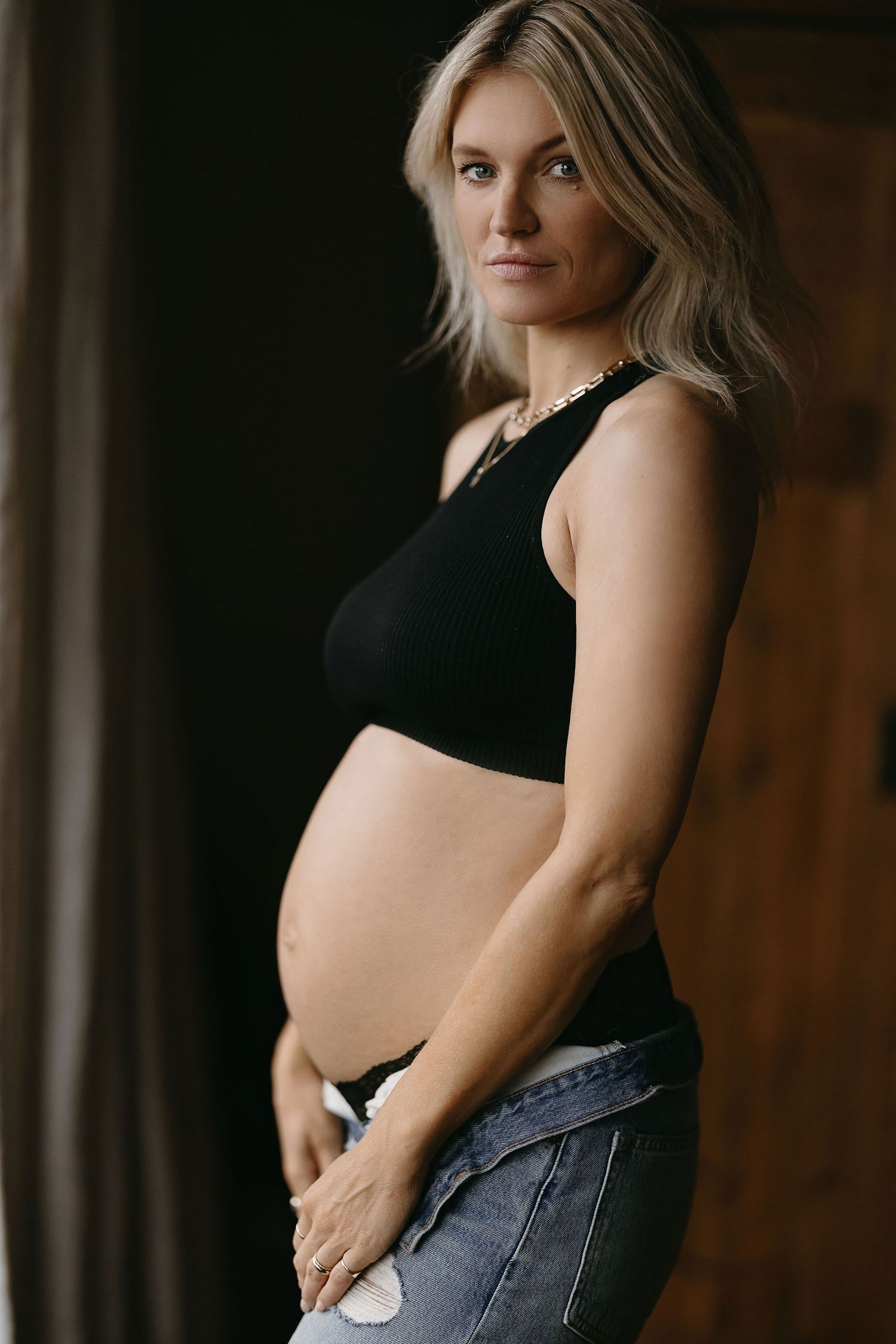 Side profile maternity photo with expectant mother in black crop top and jeans against dark background.