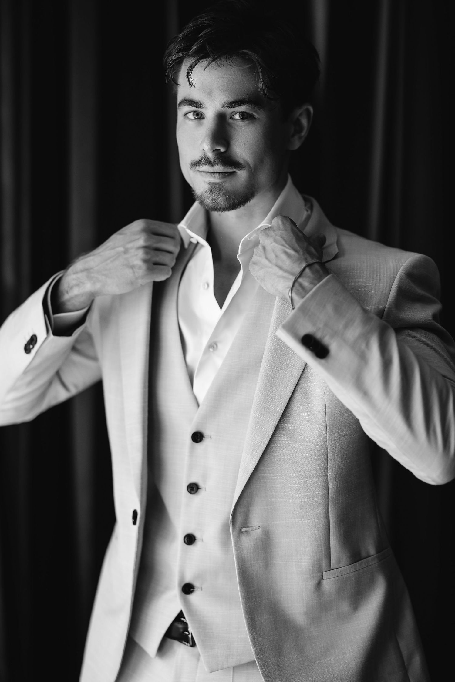 Black and white portrait of a man in a crisp white three-piece suit adjusting his collar against a dark background.