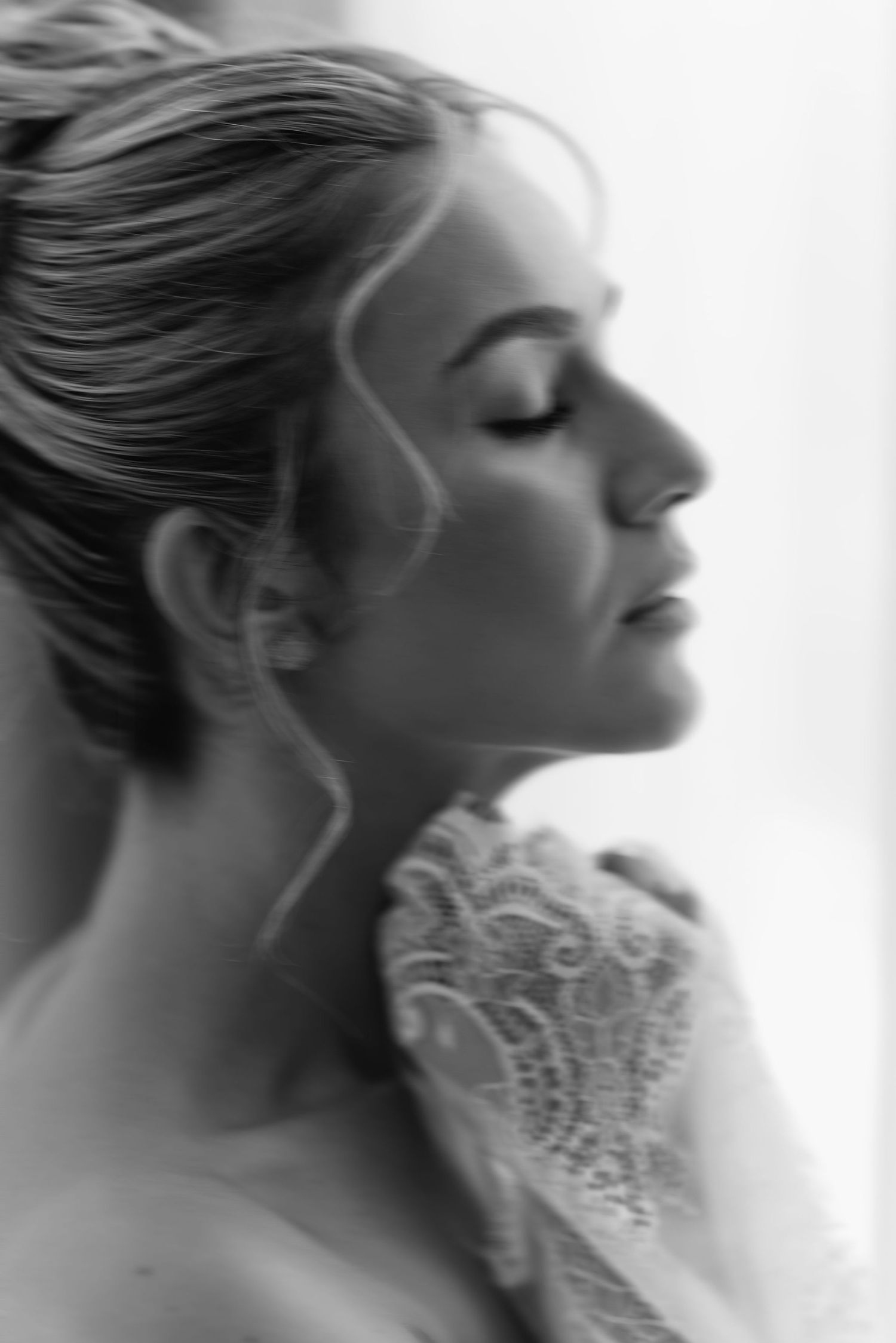 A black and white portrait in soft focus showing lace wedding dress details and swept back hair.