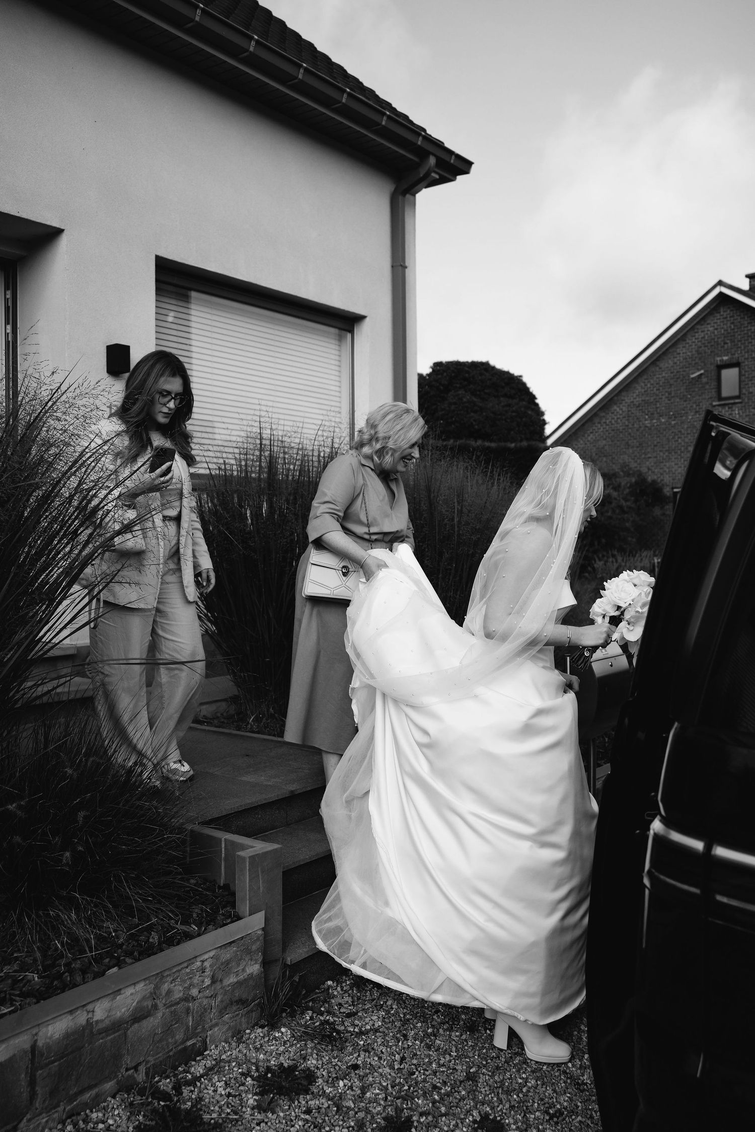 Black and white candid shot of wedding party helping bride into car outside residence.