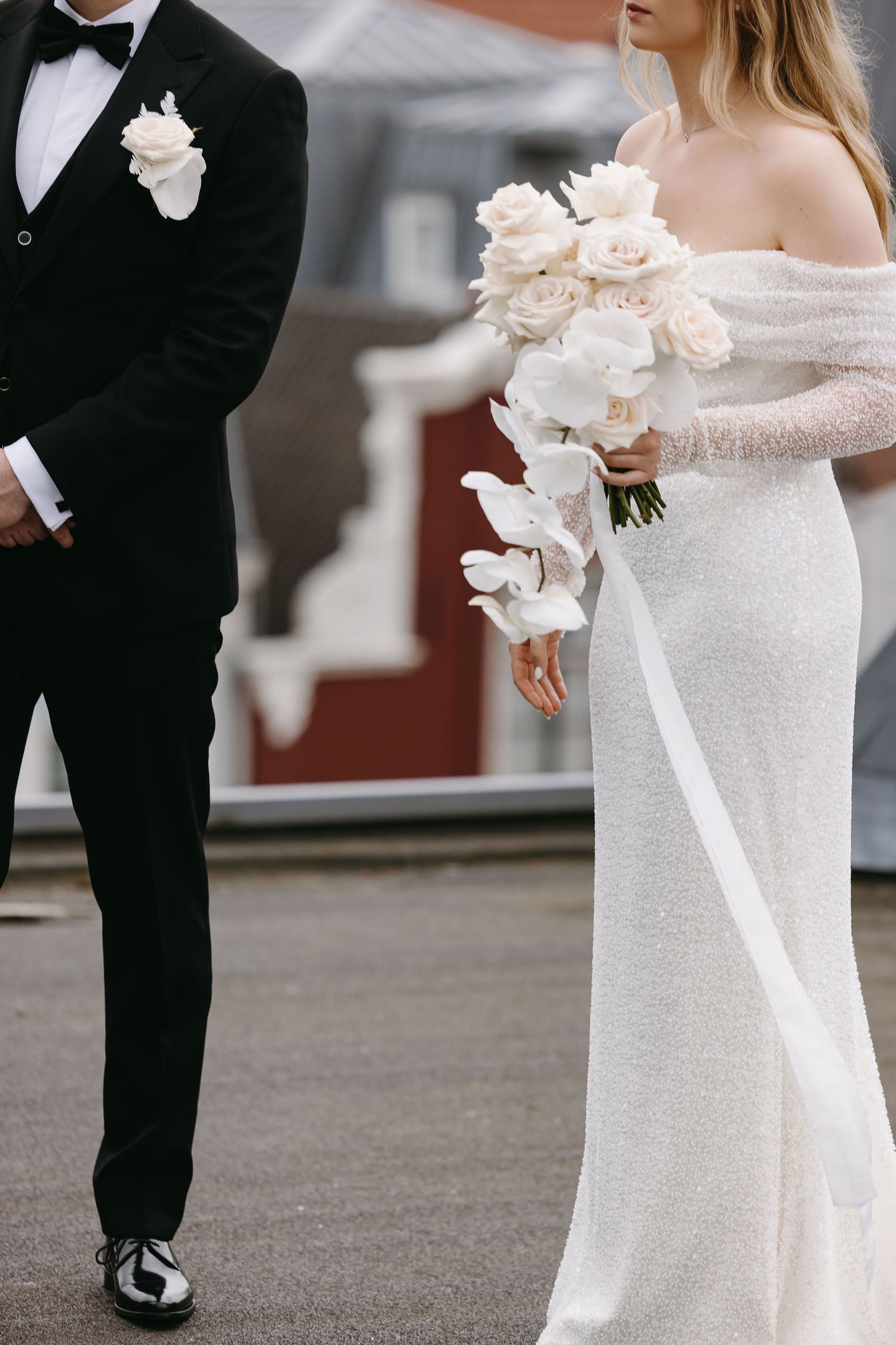 A bride in an off-shoulder gown holds white roses next to someone in a classic tuxedo.