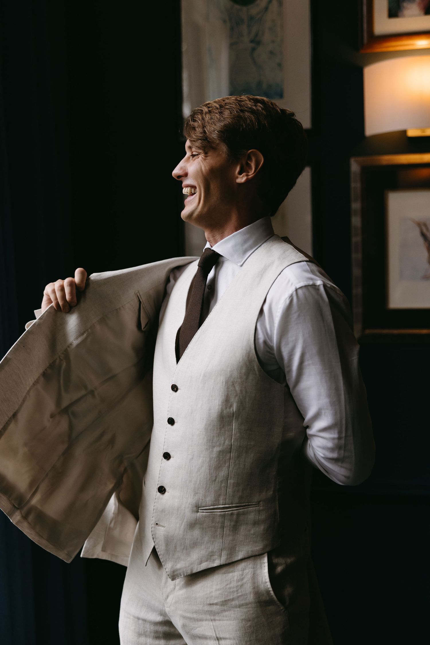 A person in a light colored suit vest and tie smiles while getting dressed in a dark room.