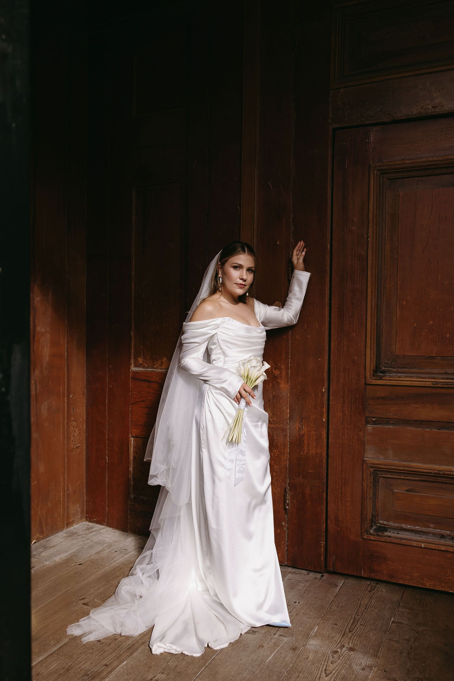 Bride in long sleeve white gown and veil sitting against wooden wall in moody lighting.