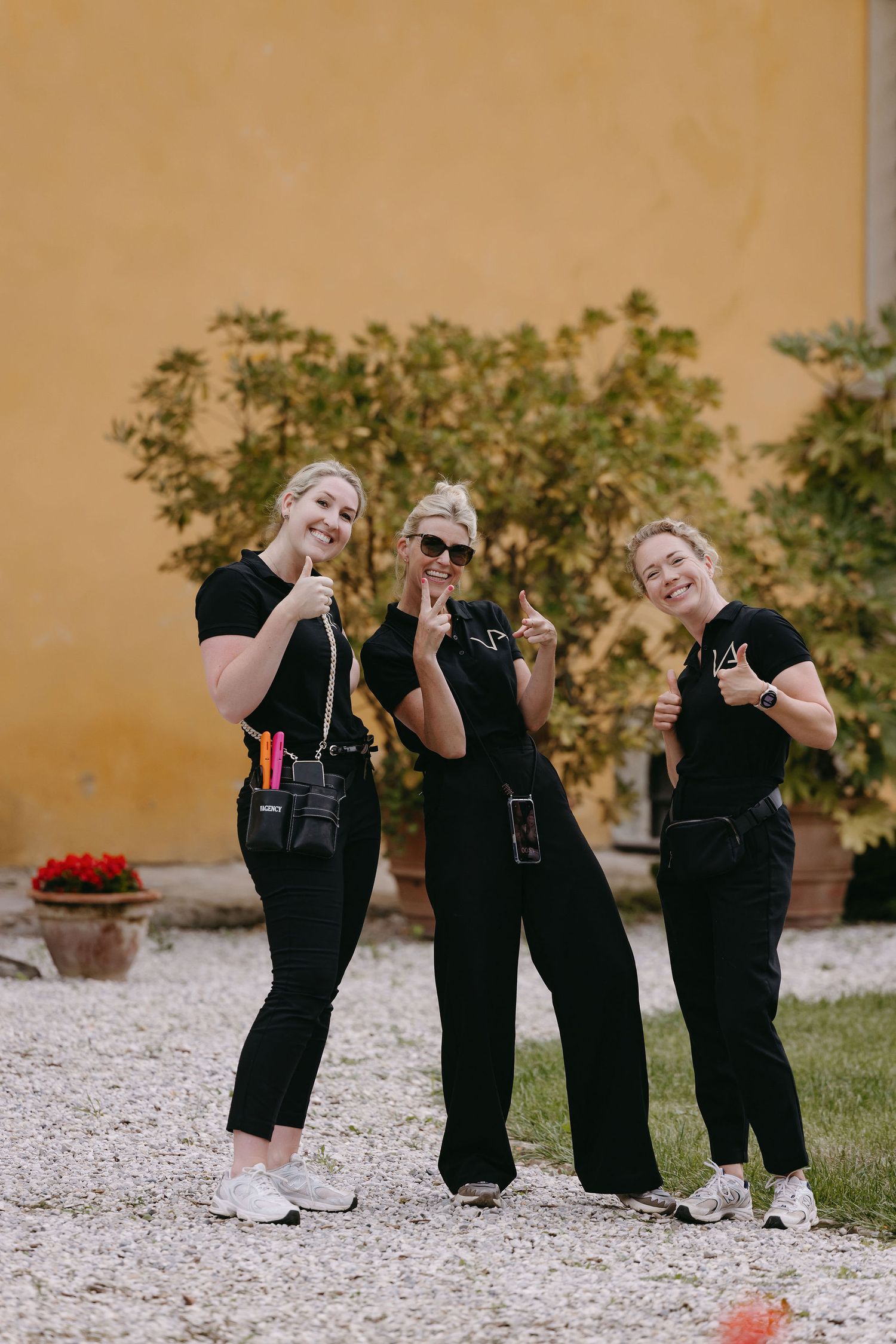 Three women in matching black outfits pose playfully against a yellow wall background.