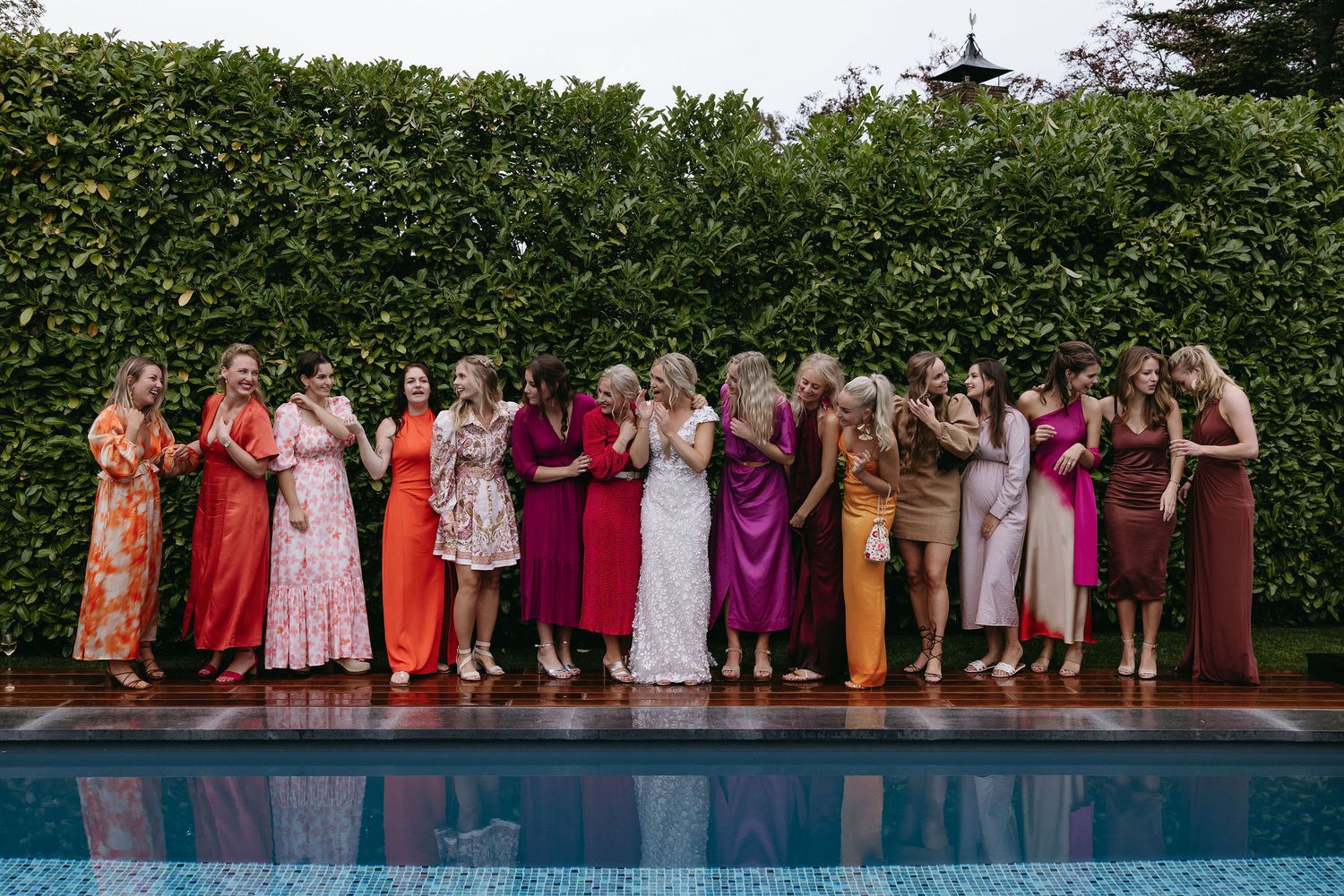 Wedding party members in colorful dresses pose together by a pool against a lush green hedge backdrop.
