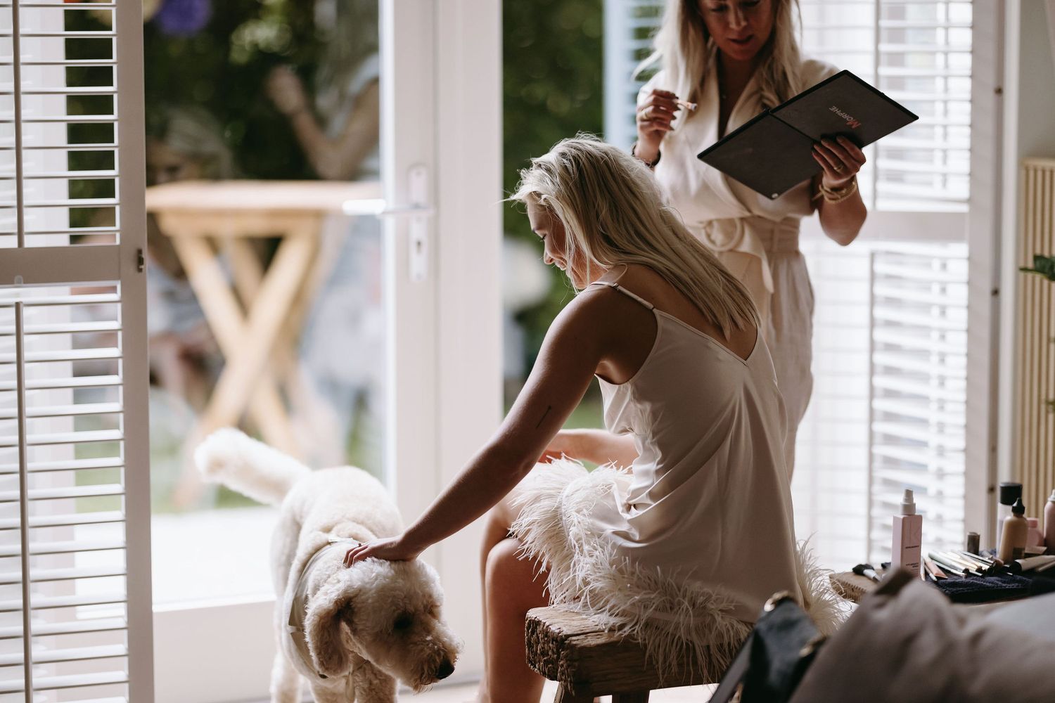 A person in white dress pets a fluffy dog while getting ready in a bright sunlit room.