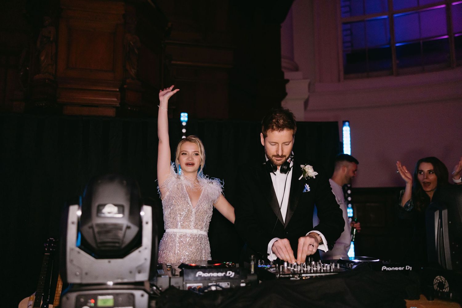 A couple celebrating at their wedding reception behind DJ equipment with dramatic purple lighting.