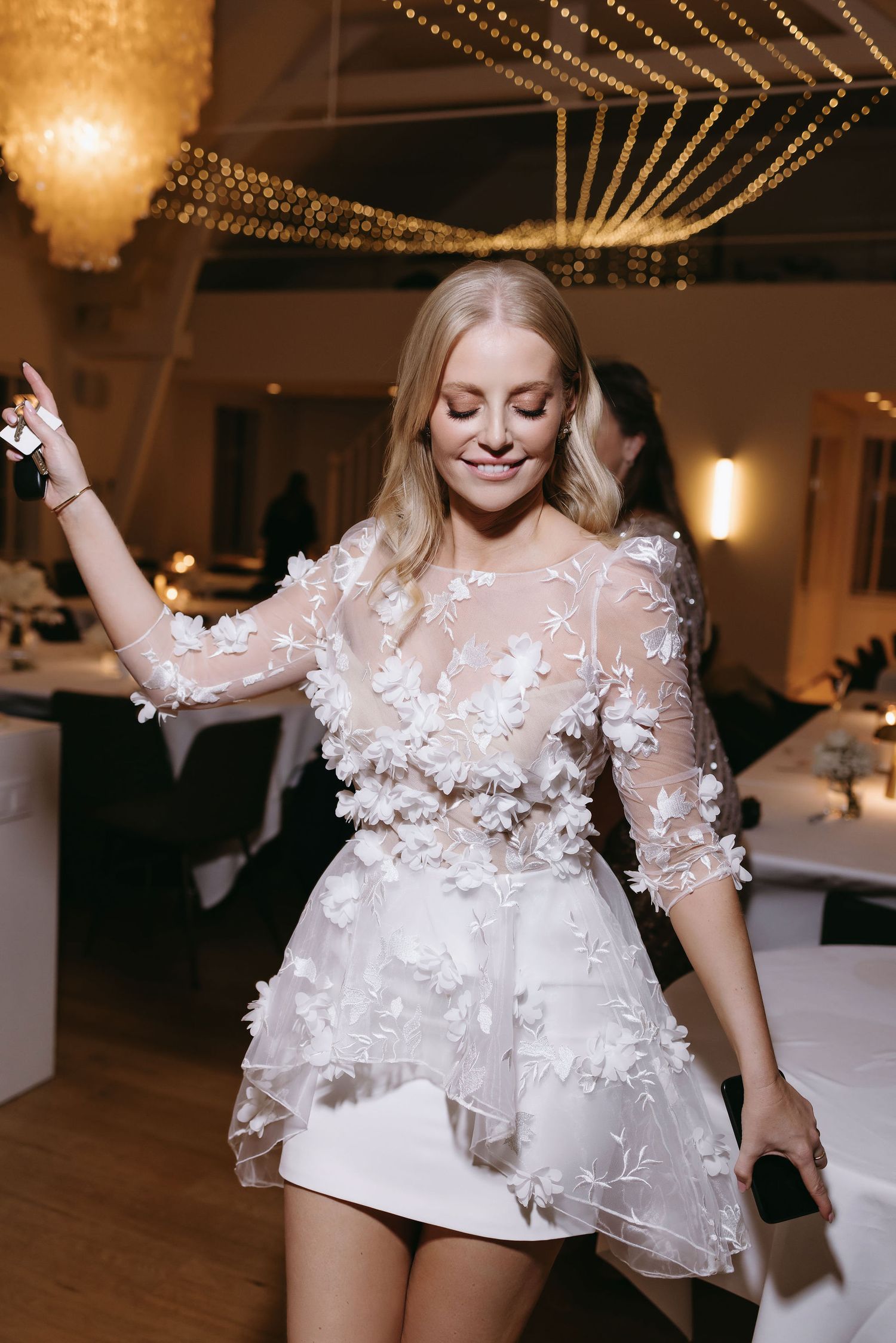 Bride dancing in short white reception dress with sheer floral applique sleeves under string lights.