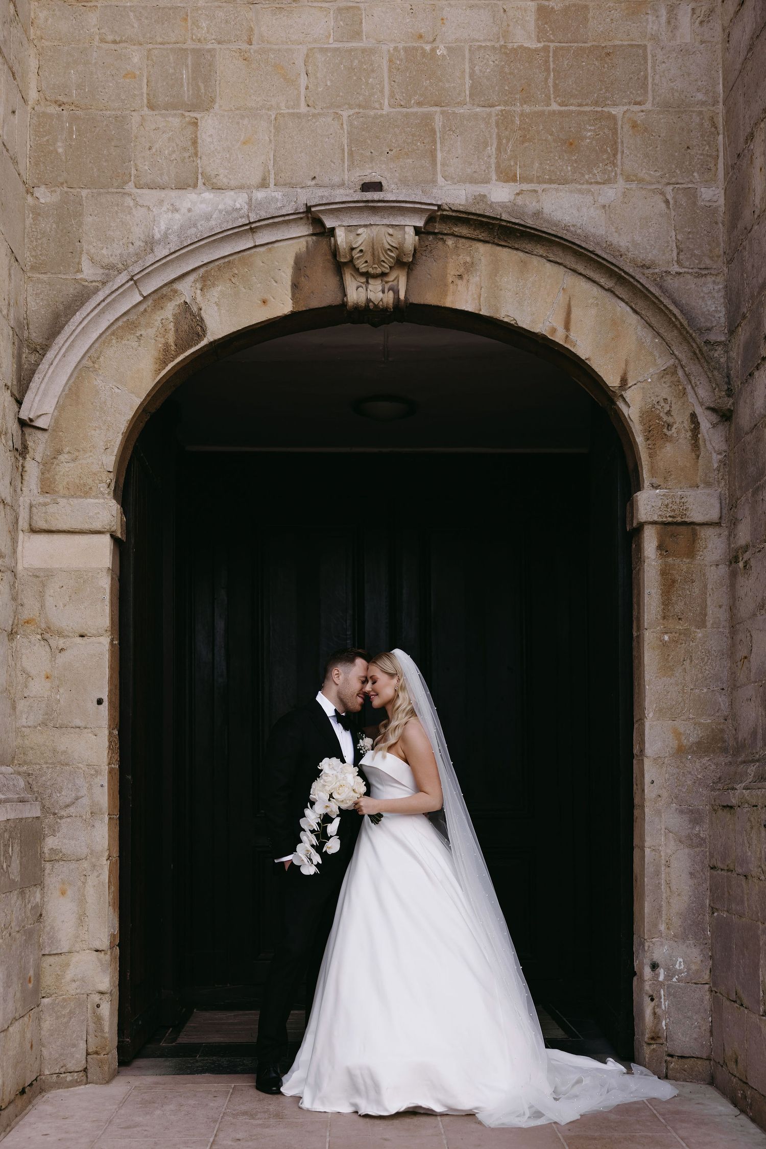 A romantic wedding couple shares an intimate moment under a stone archway in traditional formal attire.