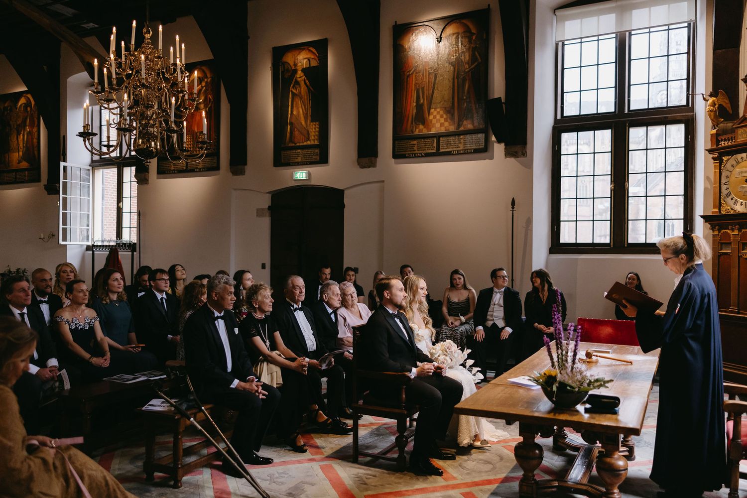 Wide shot of a traditional church ceremony with attendees seated in ornate hall under chandeliers.