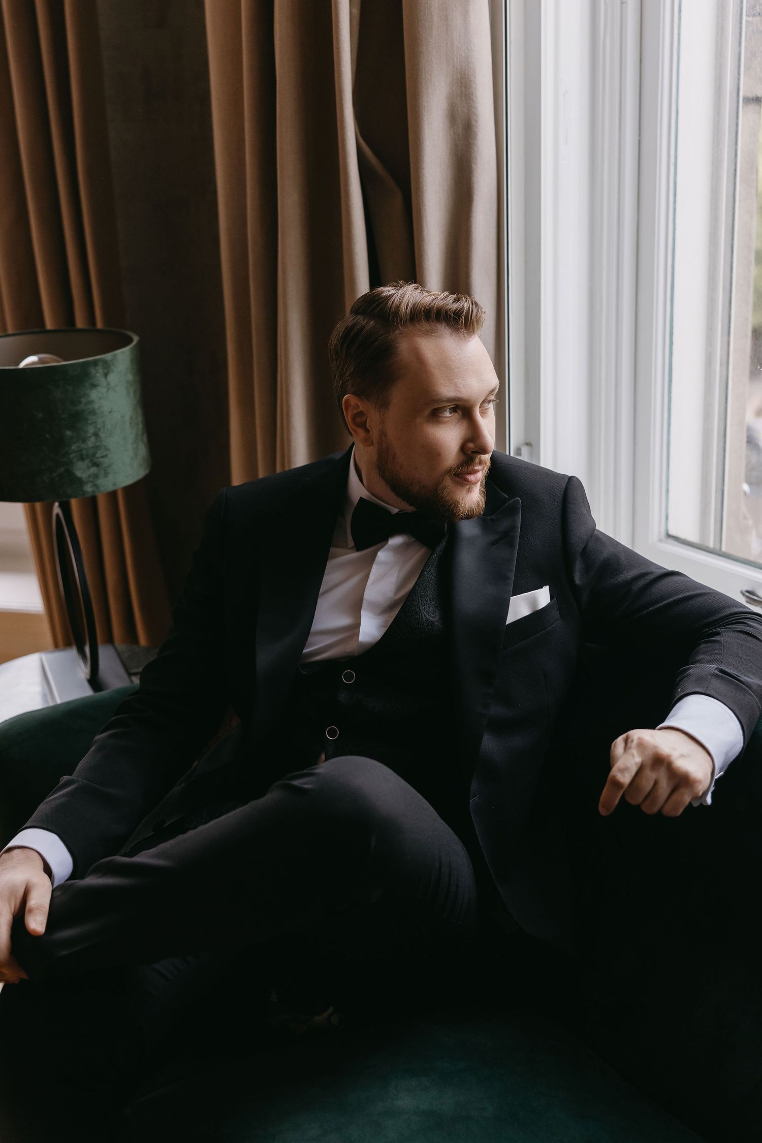 A formal portrait of someone in a black tuxedo seated on a green velvet chair by a window.