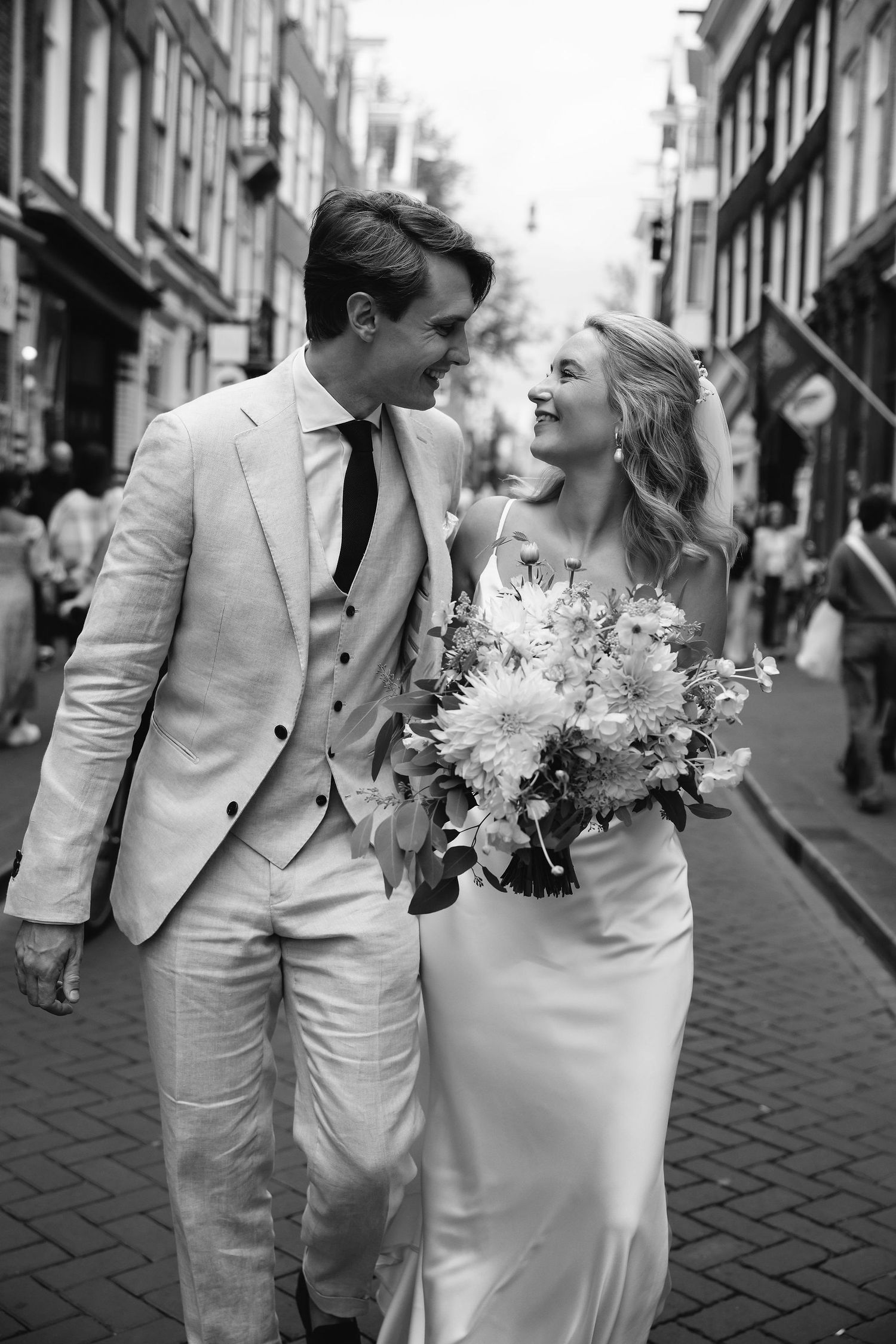 Black and white photo of couple in wedding attire walking cobblestone street with bouquet.