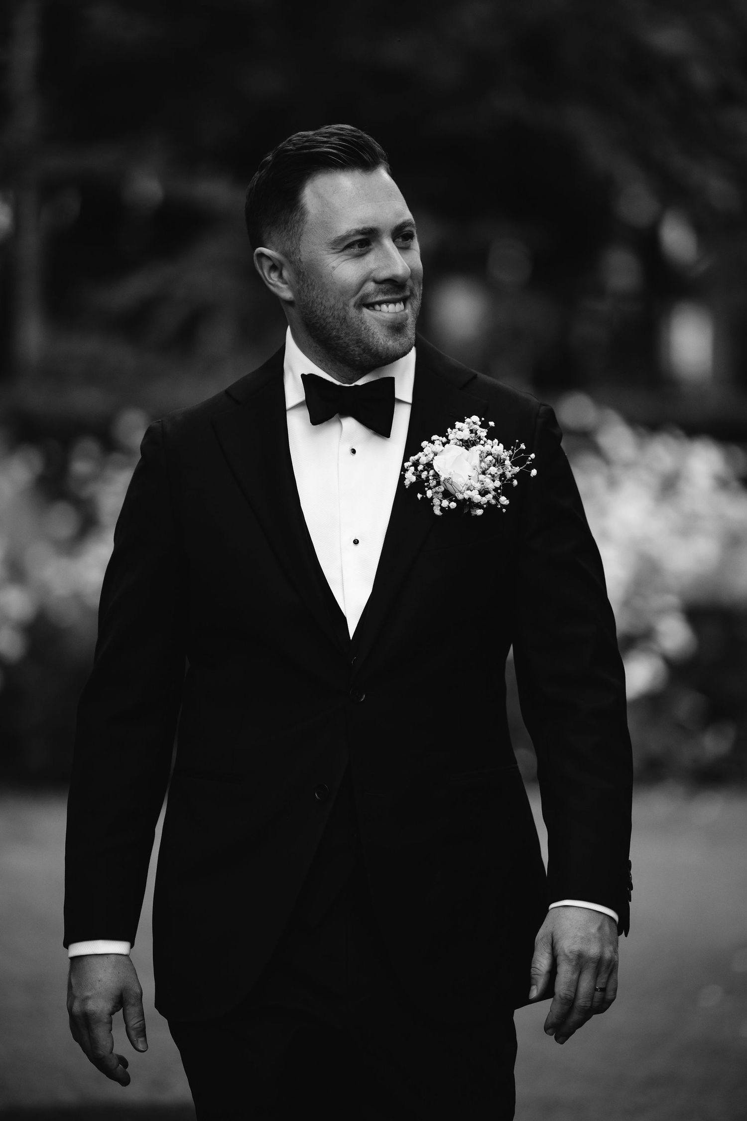 Black and white portrait of a groom in tuxedo and bow tie with a boutonniere smiling during a wedding.