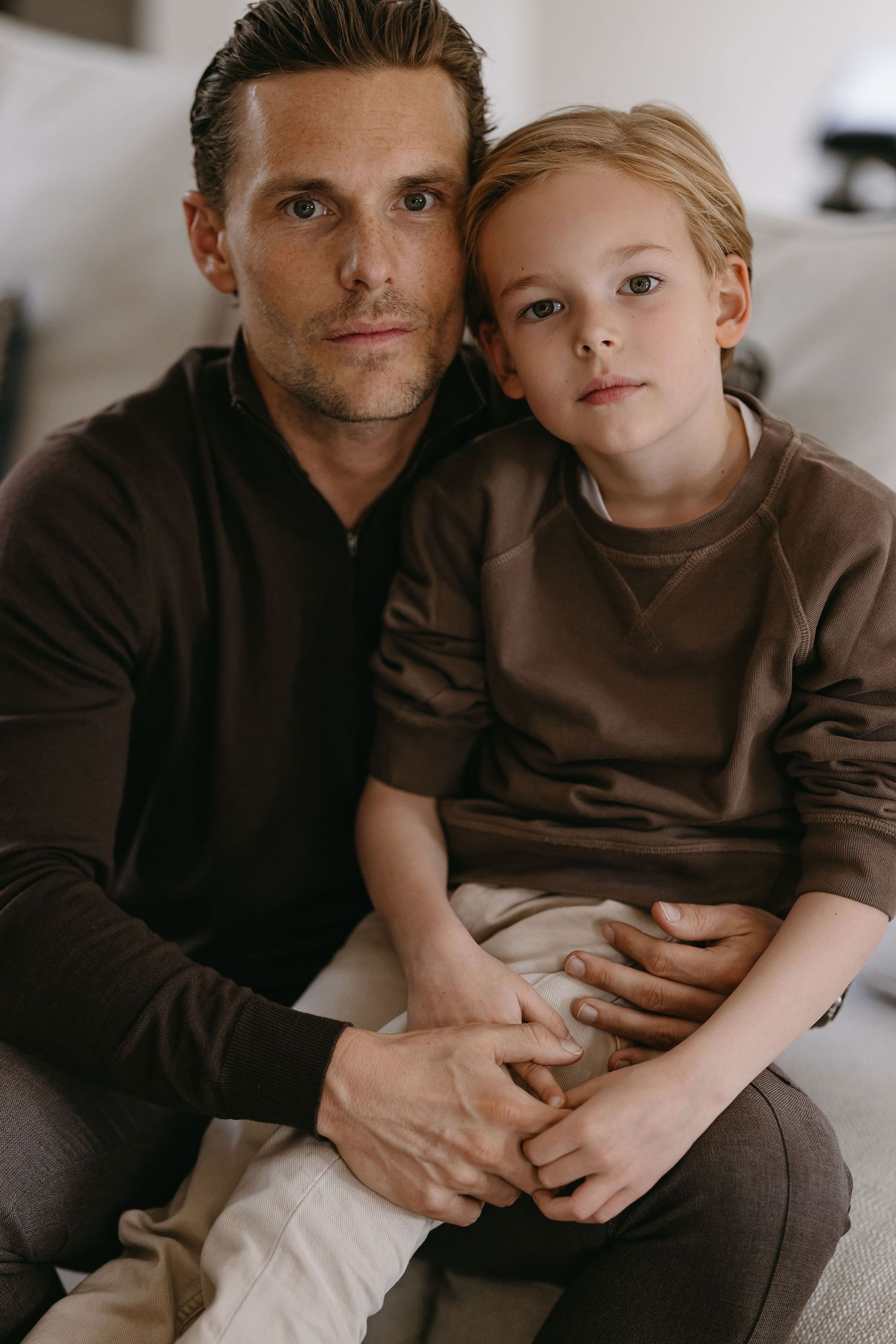 Indoor portrait of two people sitting close together on a couch, wearing casual brown clothing.