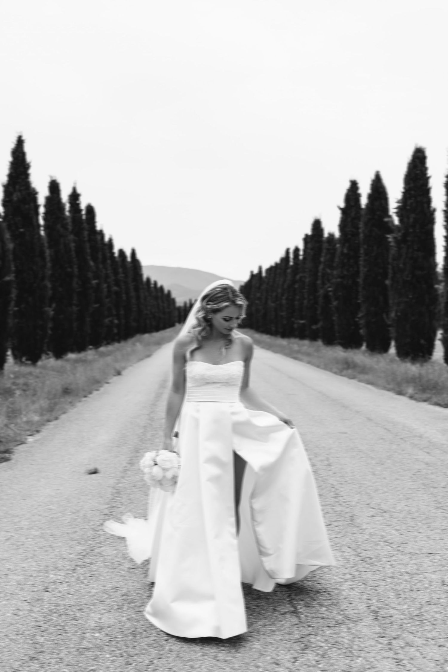 Dramatic black and white photo of a classical wedding dress on a cypress-lined road in Italy.