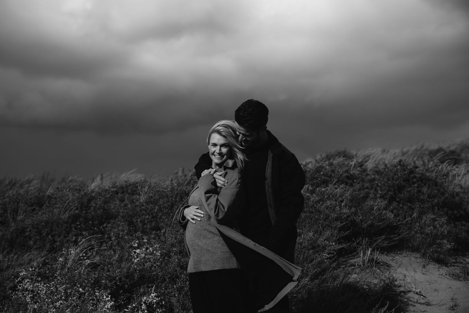 Dramatic black and white photo of couple embracing on coastal path under stormy sky.