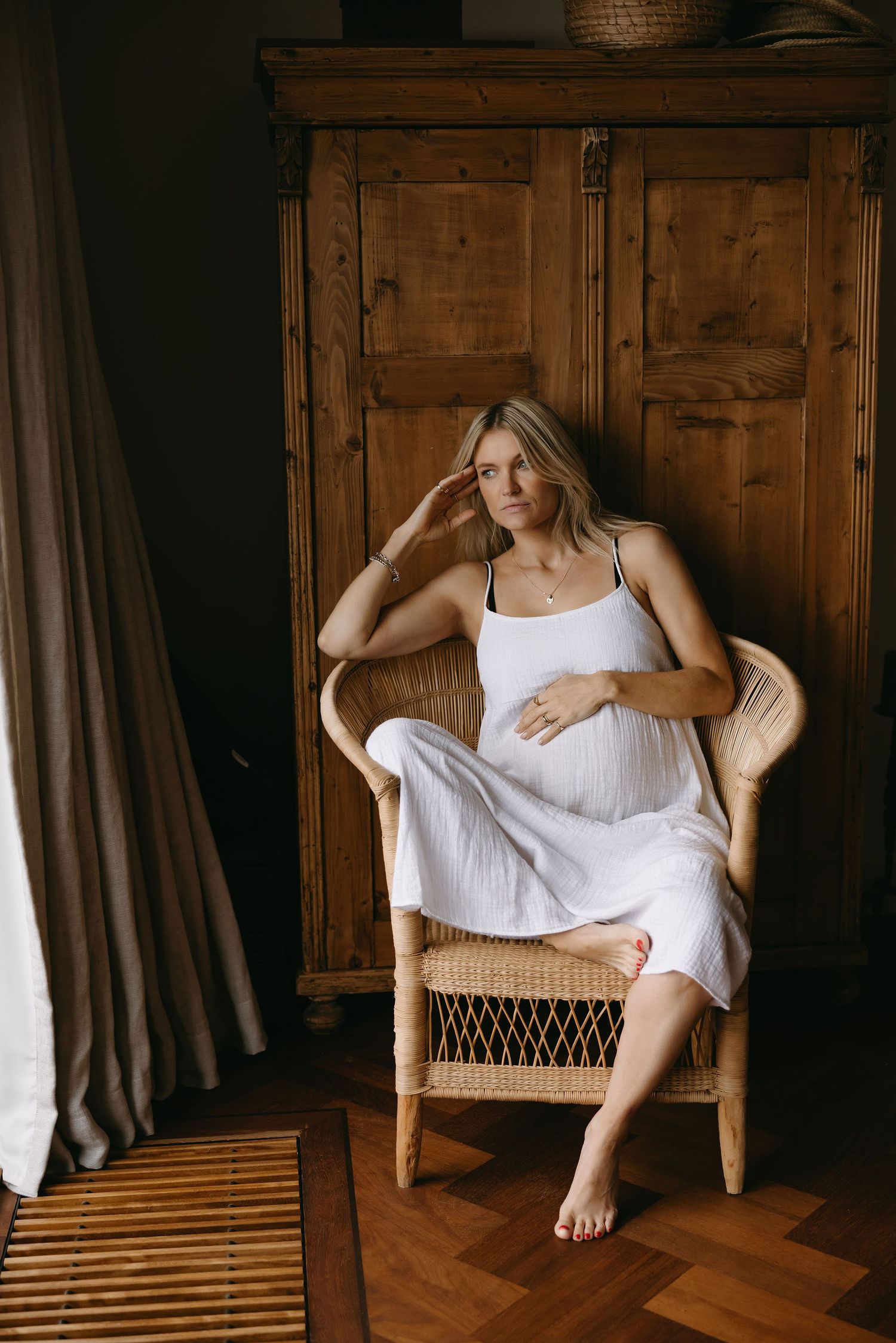 Maternity portrait of someone in white dress sitting in wicker chair by wooden wardrobe.