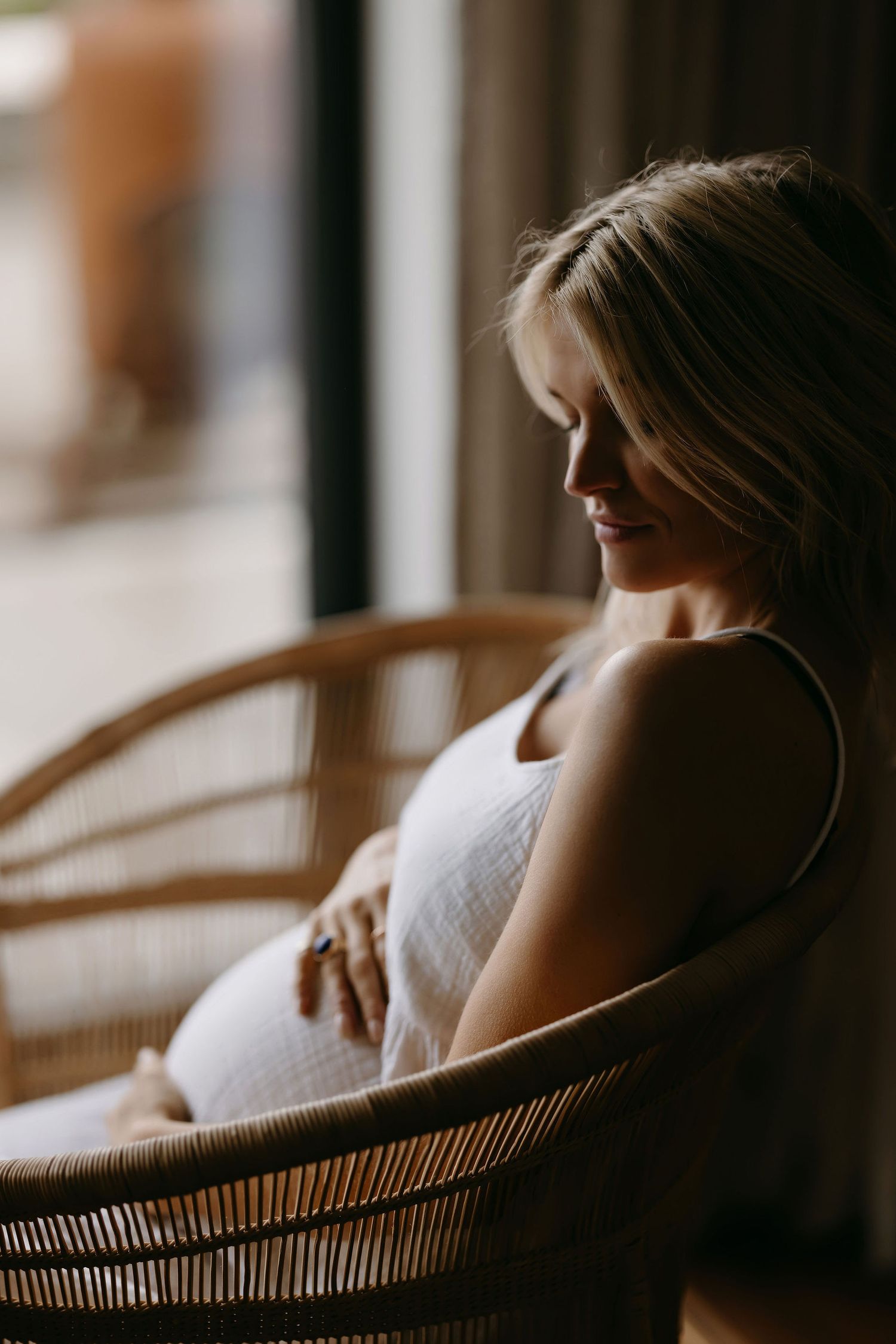 Serene maternity portrait in rattan chair with soft window lighting.