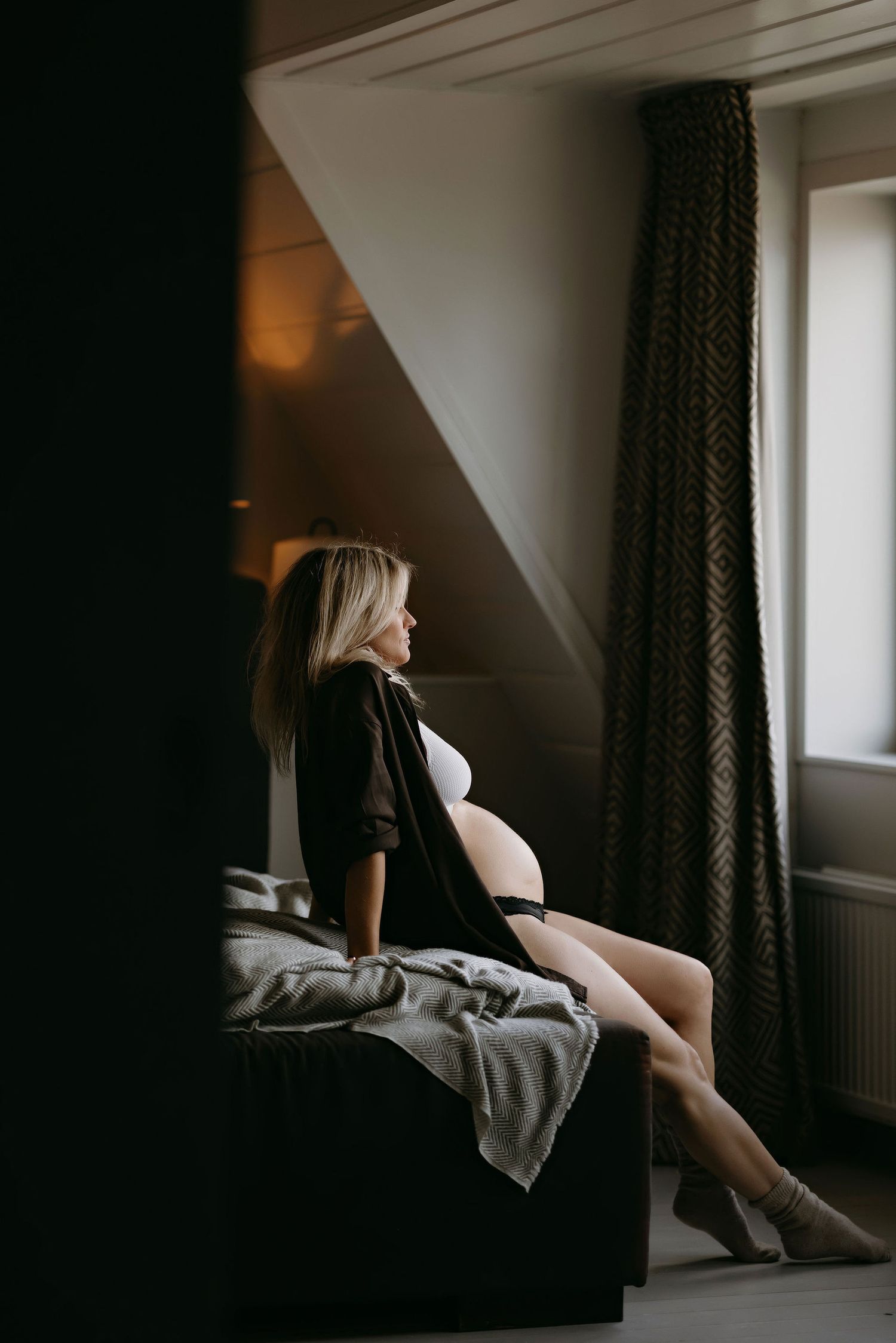 Artistic maternity portrait with mother-to-be sitting by window in dark dress.