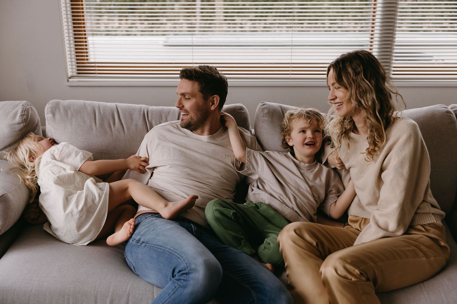 A relaxed family lounging together on a grey couch near a window.