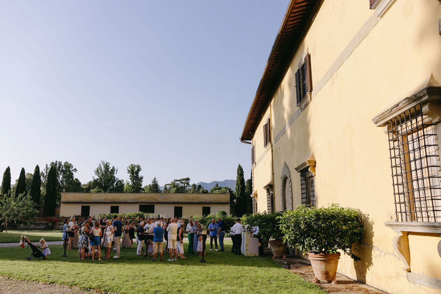 A group gathers outside an elegant Tuscan villa with cypress trees in the background.