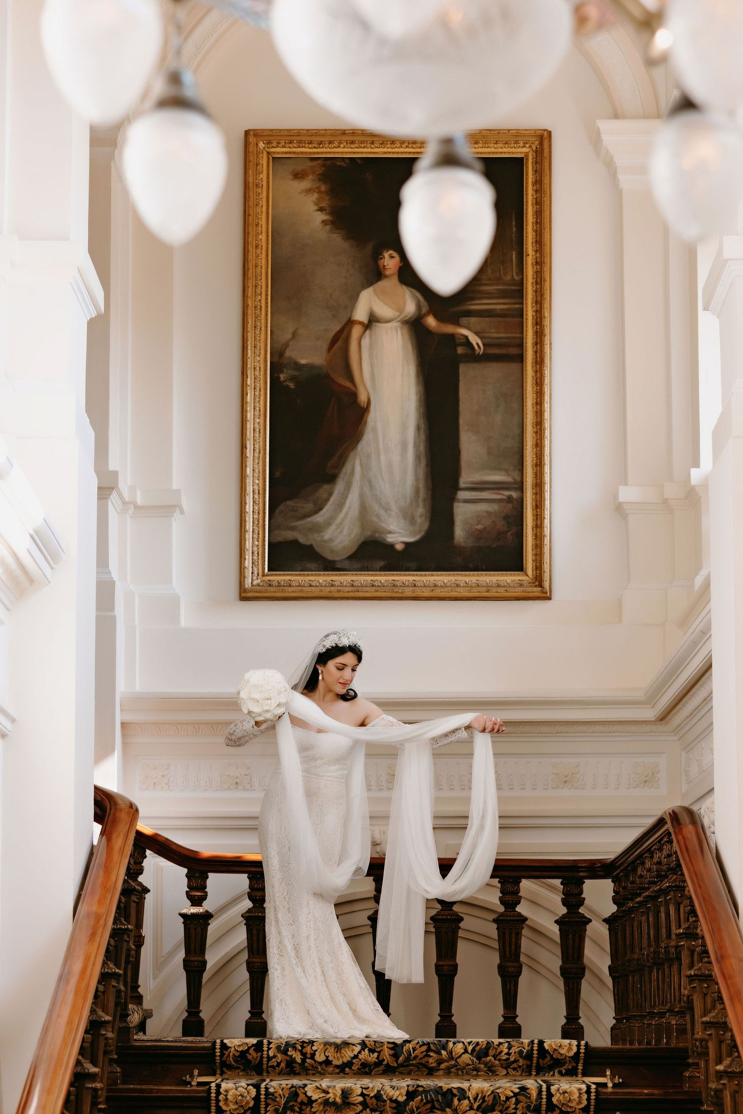 A flowing white veil drapes elegantly down a wooden staircase with a classic portrait painting displayed on the wall.