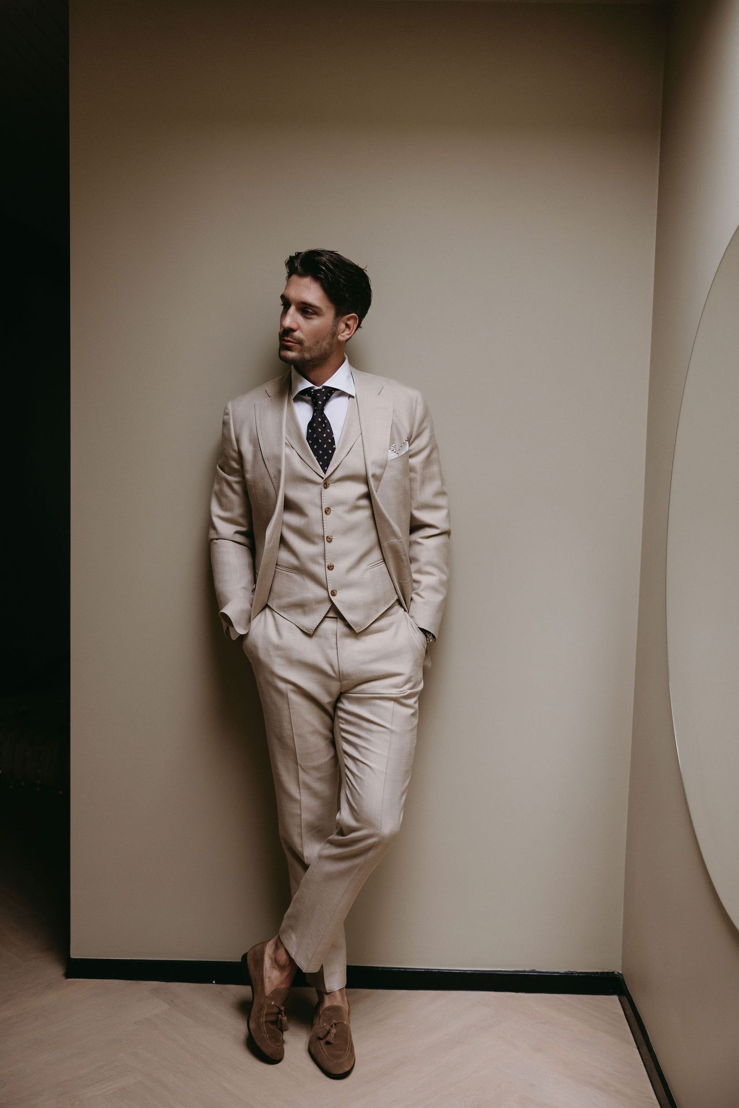 Stylish groom in a cream-colored three-piece suit and brown shoes poses against a neutral wall.