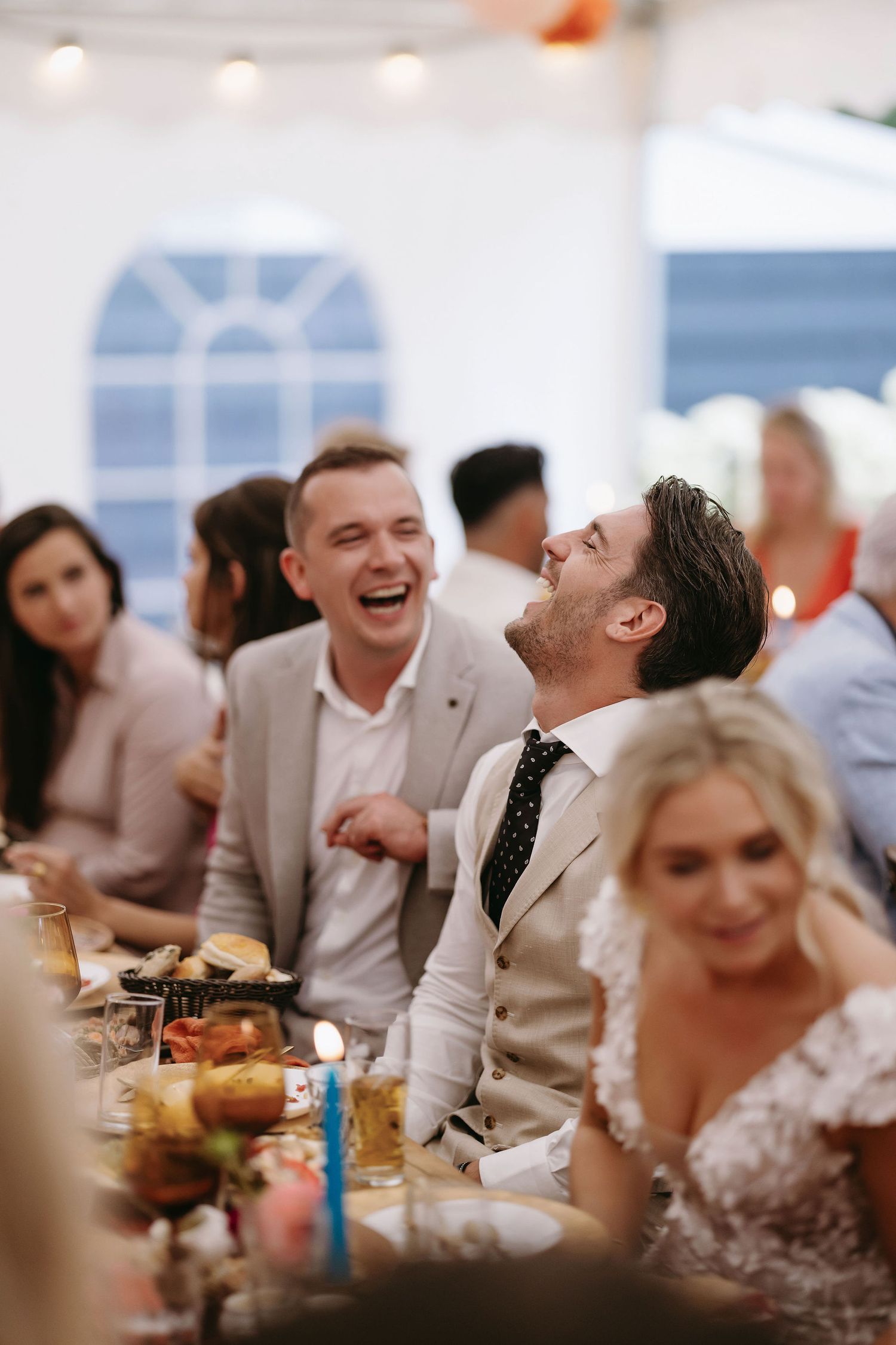 Wedding reception guests share laughter and conversation at tables under a white tent.