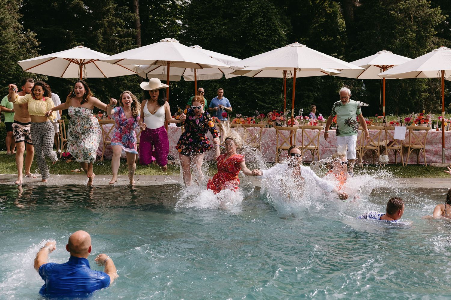 Joyful pool party scene with people splashing in the water under white umbrellas.