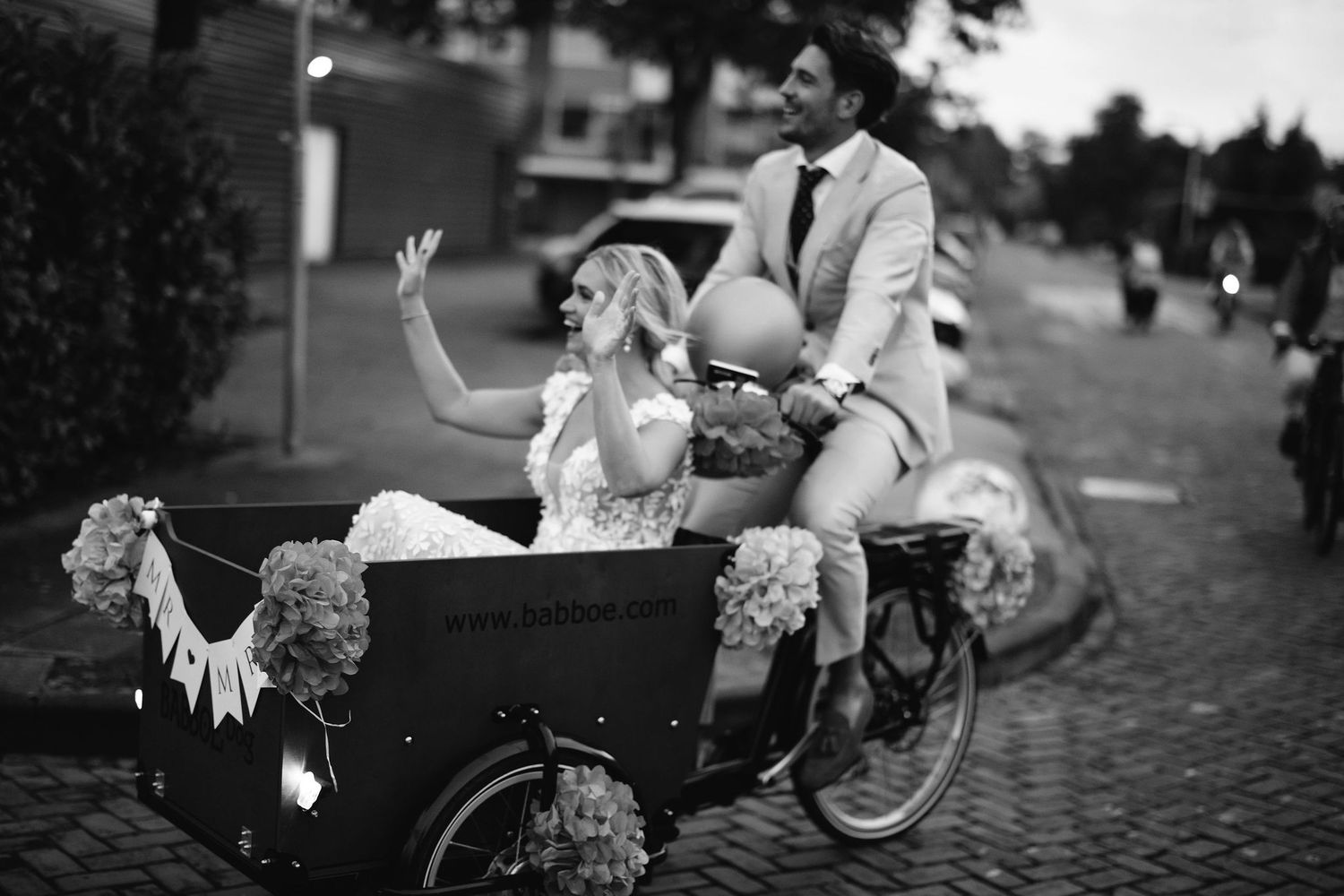 Black and white wedding photo of couple riding decorated cargo bike.