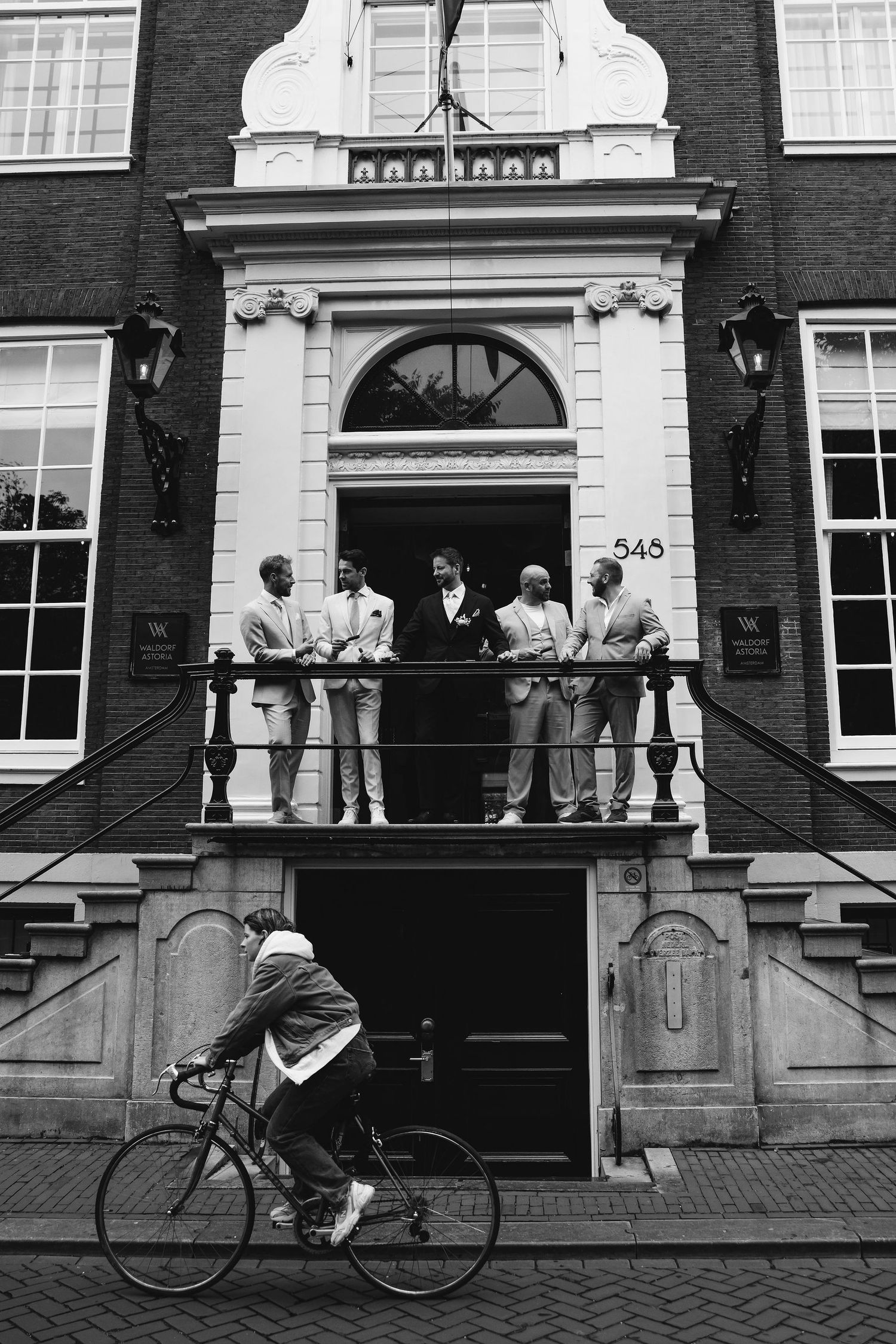 People gathered on a historic building's balcony while a cyclist rides past below.