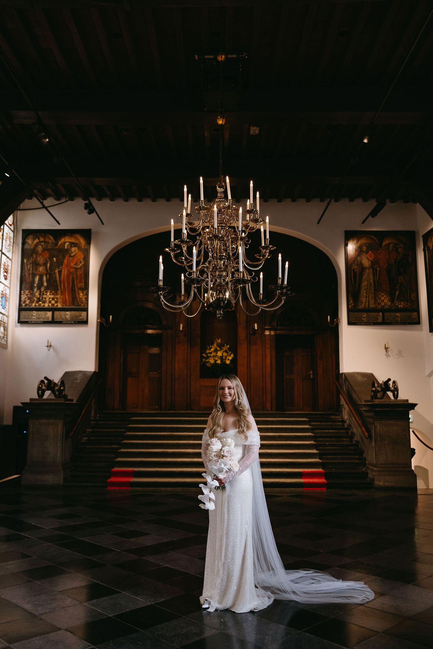 Elegant bride in long white dress stands beneath ornate chandelier in grand historic hall with paintings and ornate details.
