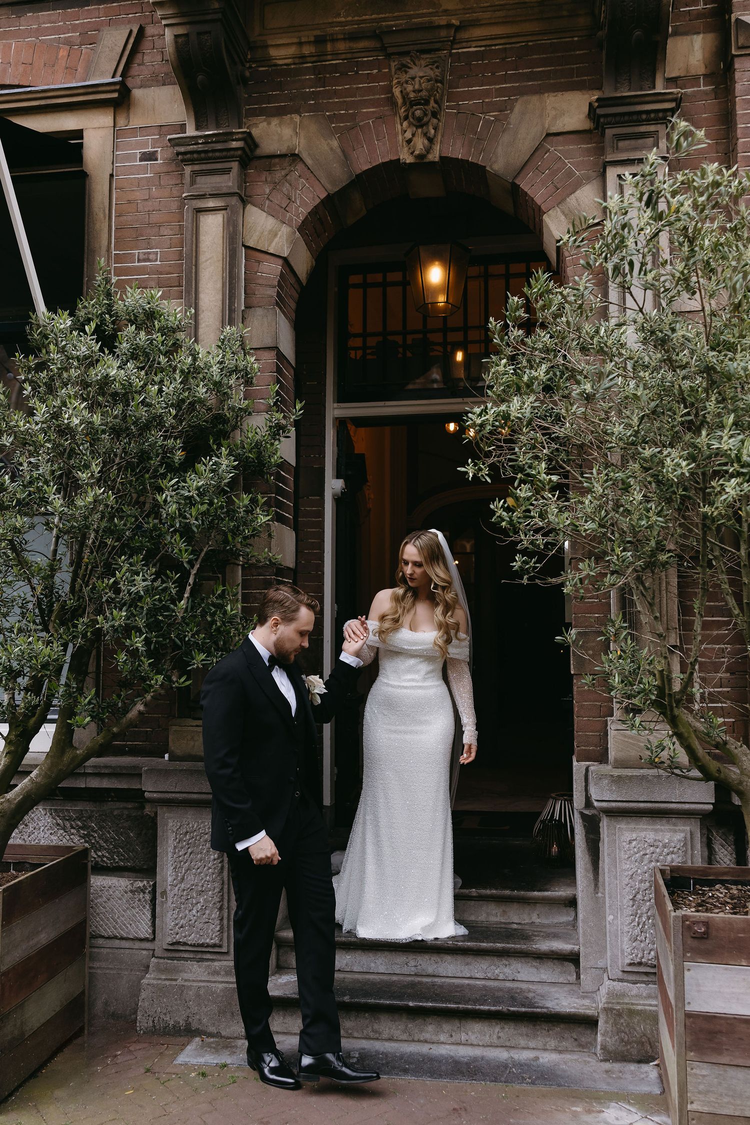 Wedding couple stands on stone steps of historic brownstone building with potted trees.