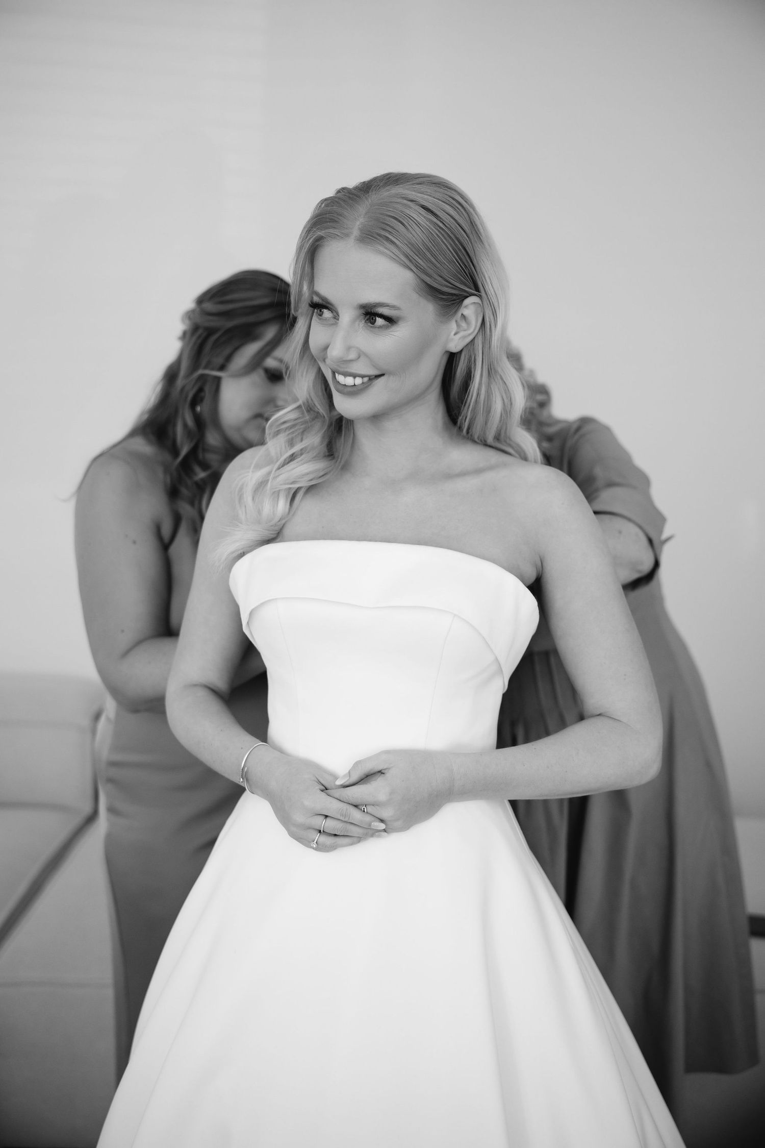 Getting ready moments with attendants helping adjust a strapless white wedding gown.