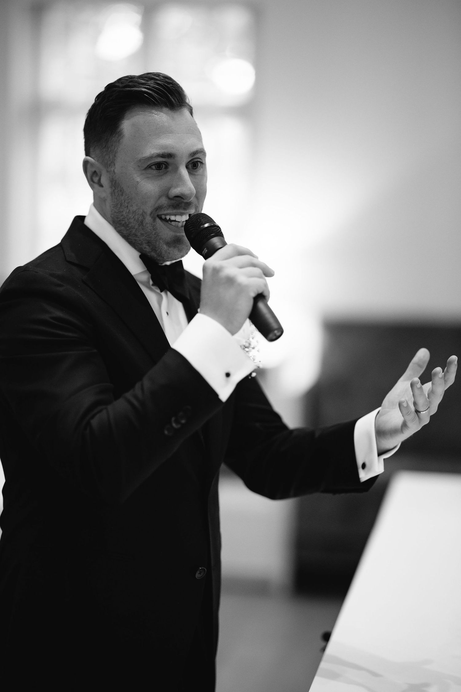 Black and white portrait of performer in tuxedo energetically speaking into microphone.
