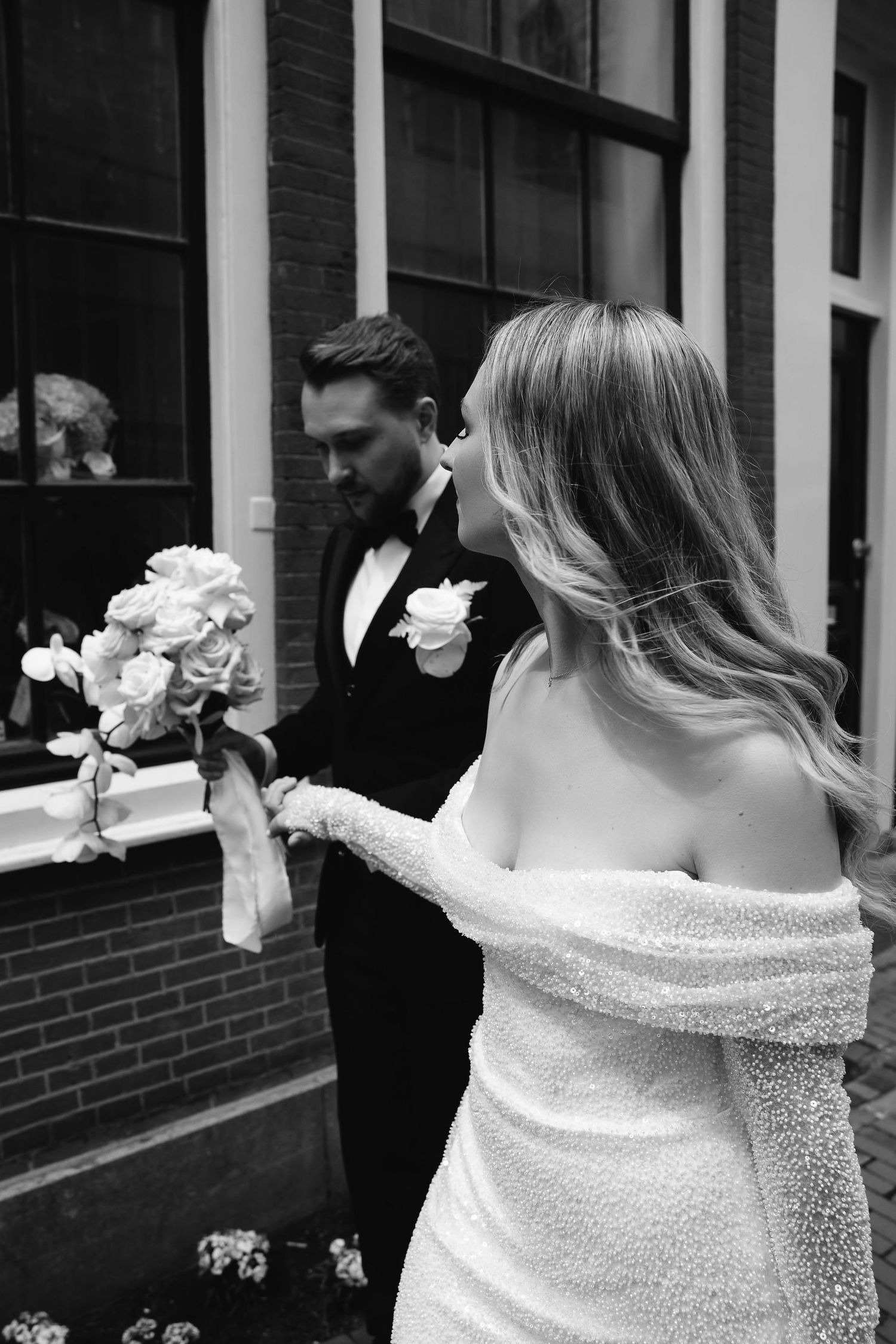 An intimate black and white wedding photo showing a couple in formal attire with white floral bouquet against a brick wall.