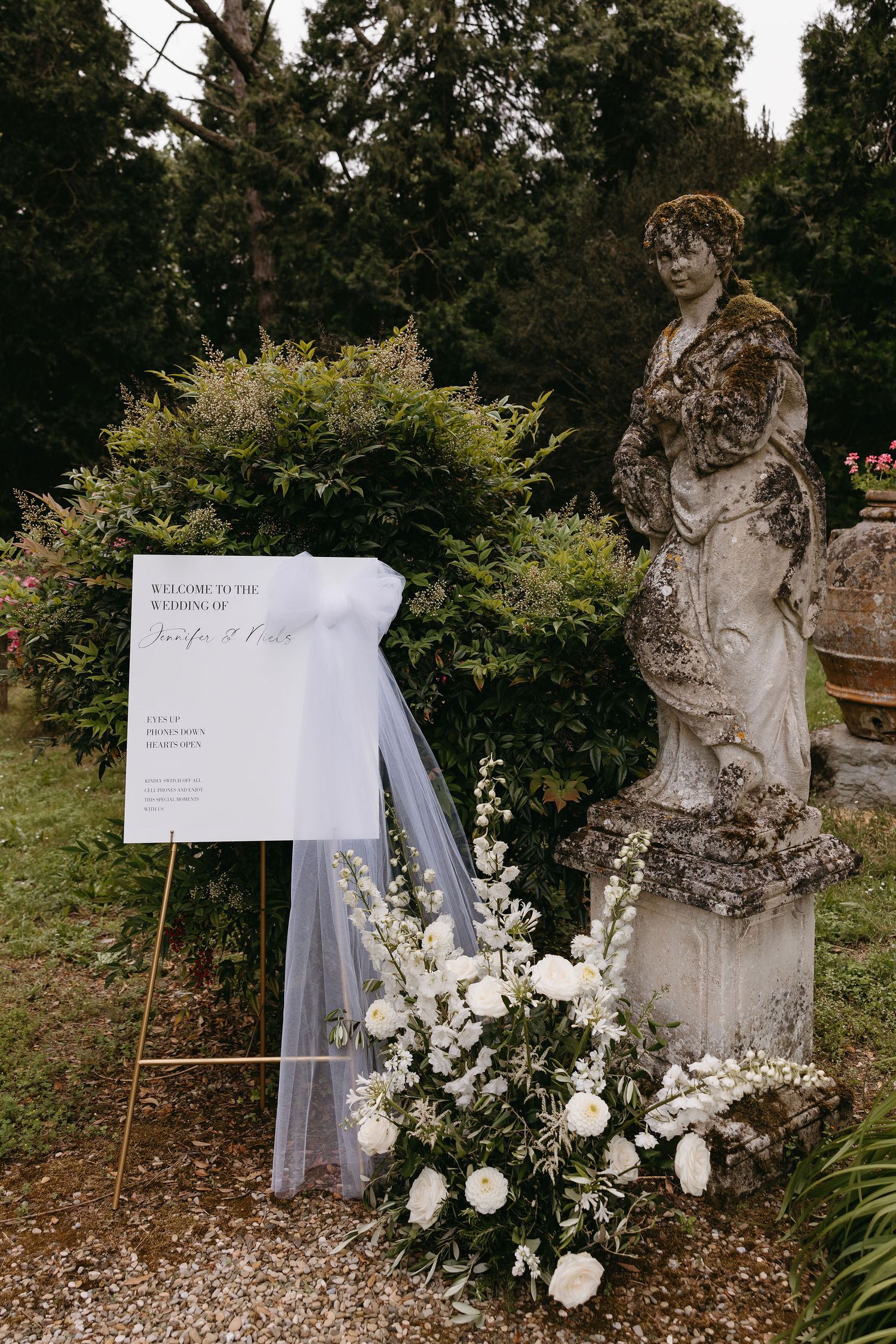 Wedding seating chart and veil displayed next to garden statue with white floral arrangement.