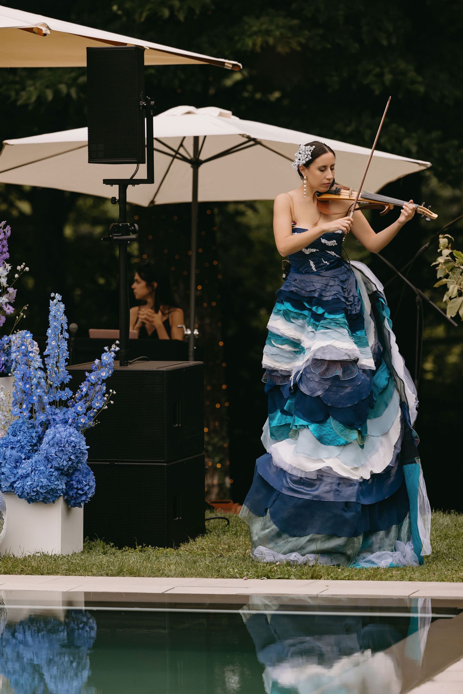 Violinist in a flowing blue gown performs poolside at an outdoor event.