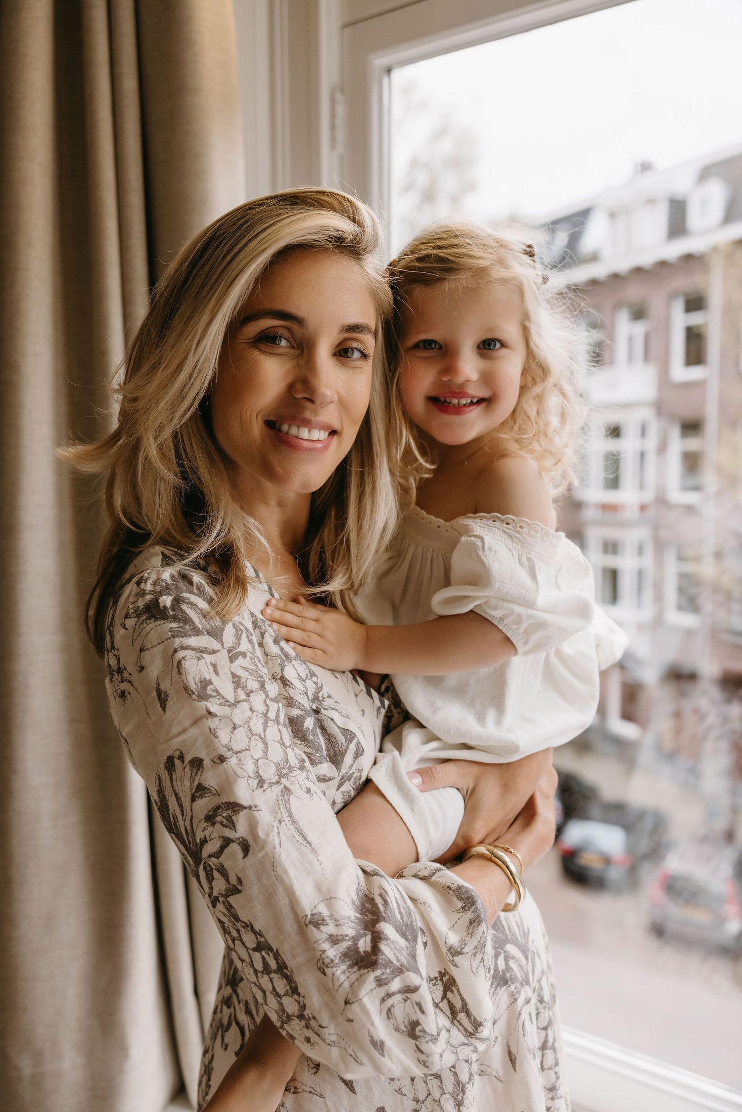 Joyful mother and daughter portrait by window in floral dress.